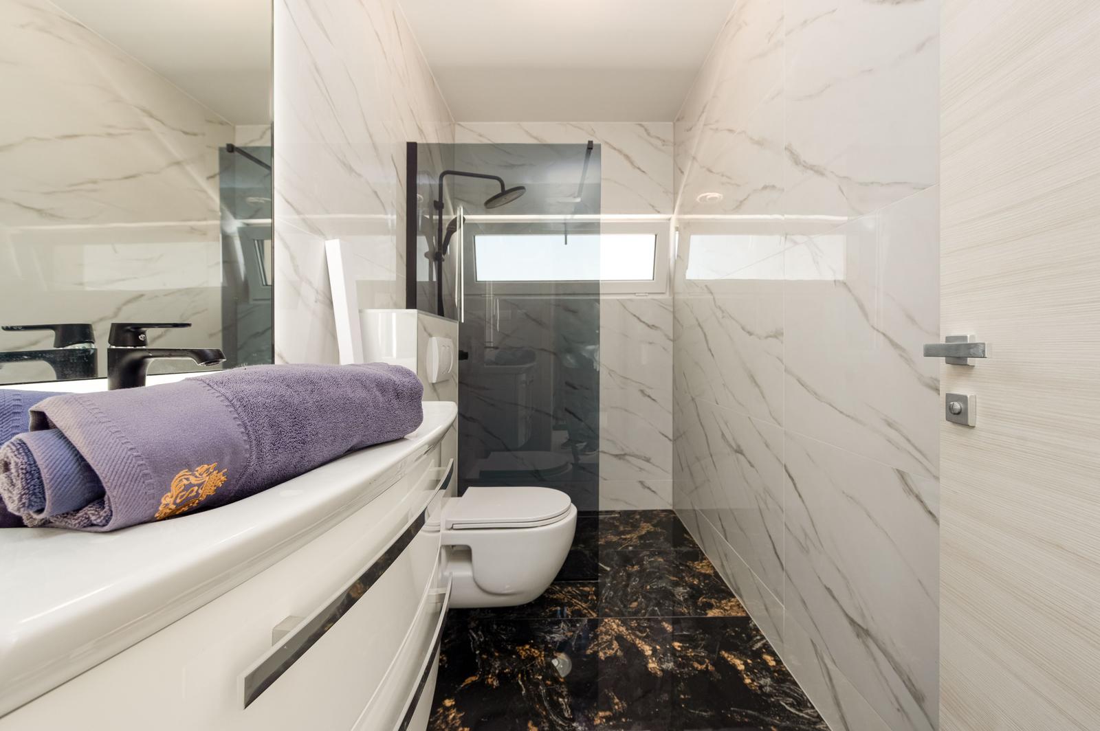Badezimmer mit Marmorwänden, Dusche, Toilette und Waschtisch mit Handtuch.