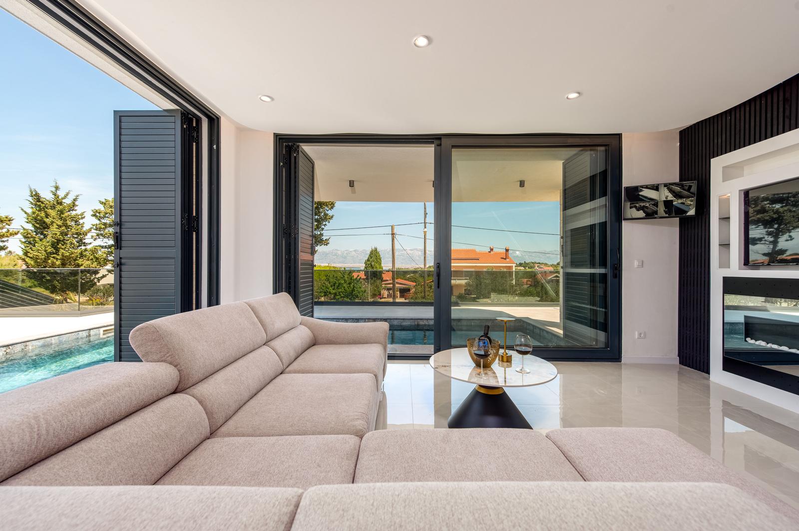 Ein Wohnbereich mit Sofa, Tisch und Fernseher. Durchgehende Glaswände öffnen sich zu einer Poolterrasse.