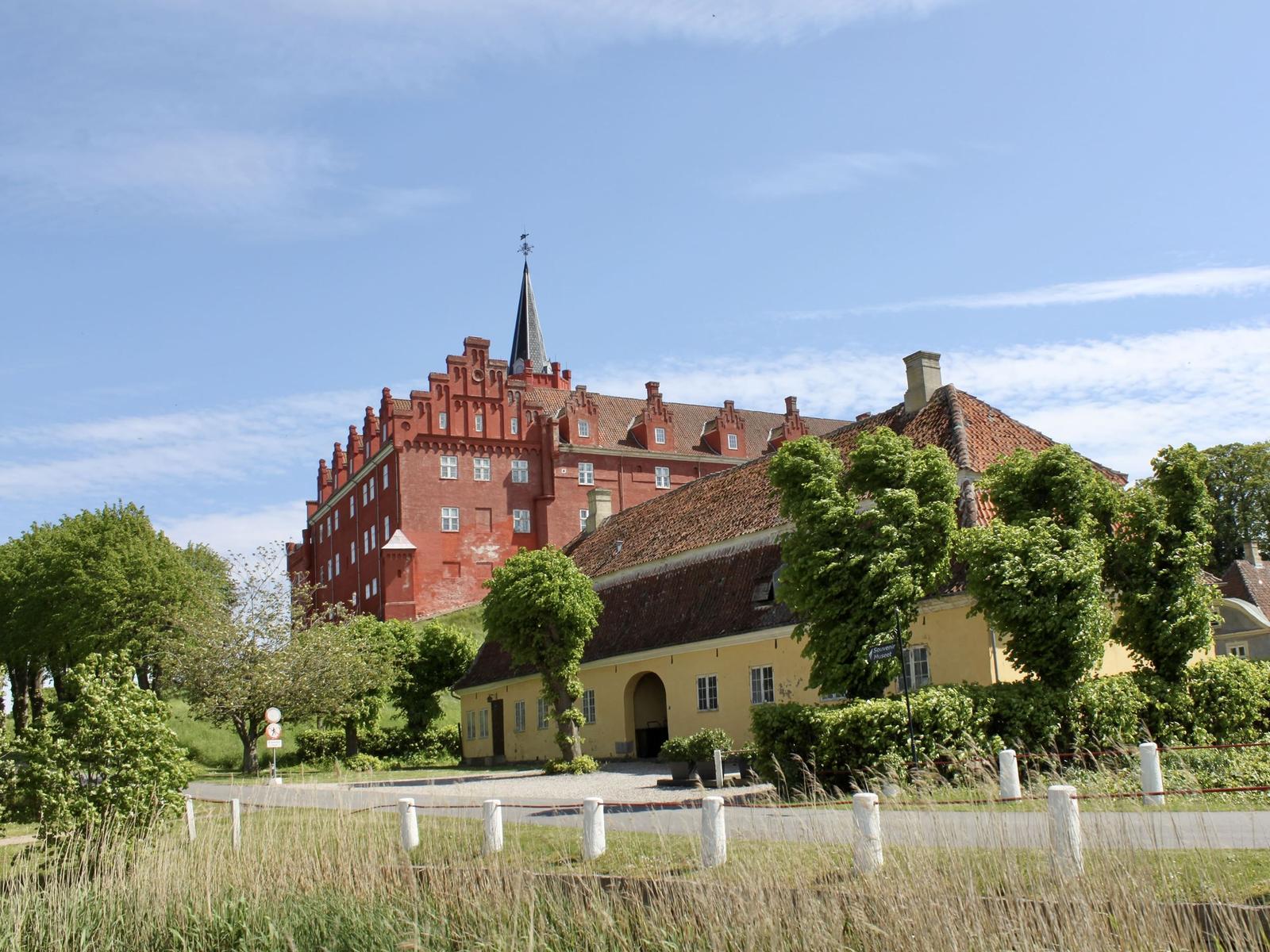 Rotes Schloss mit Turm und gelbem Nebengebäude unter blauem Himmel.