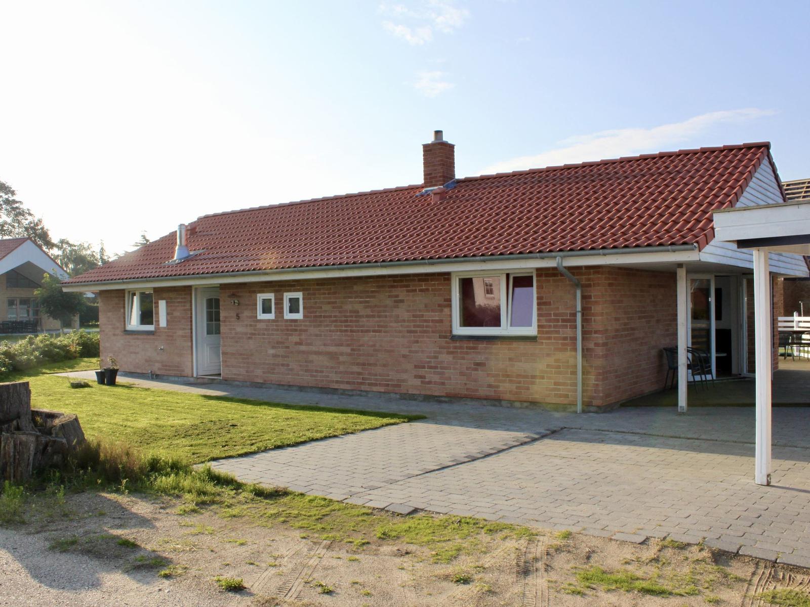 Ein eingeschossiges Haus mit Backsteinfassade und rotem Dach. Vor dem Haus befindet sich eine Terrasse und ein Rasen.