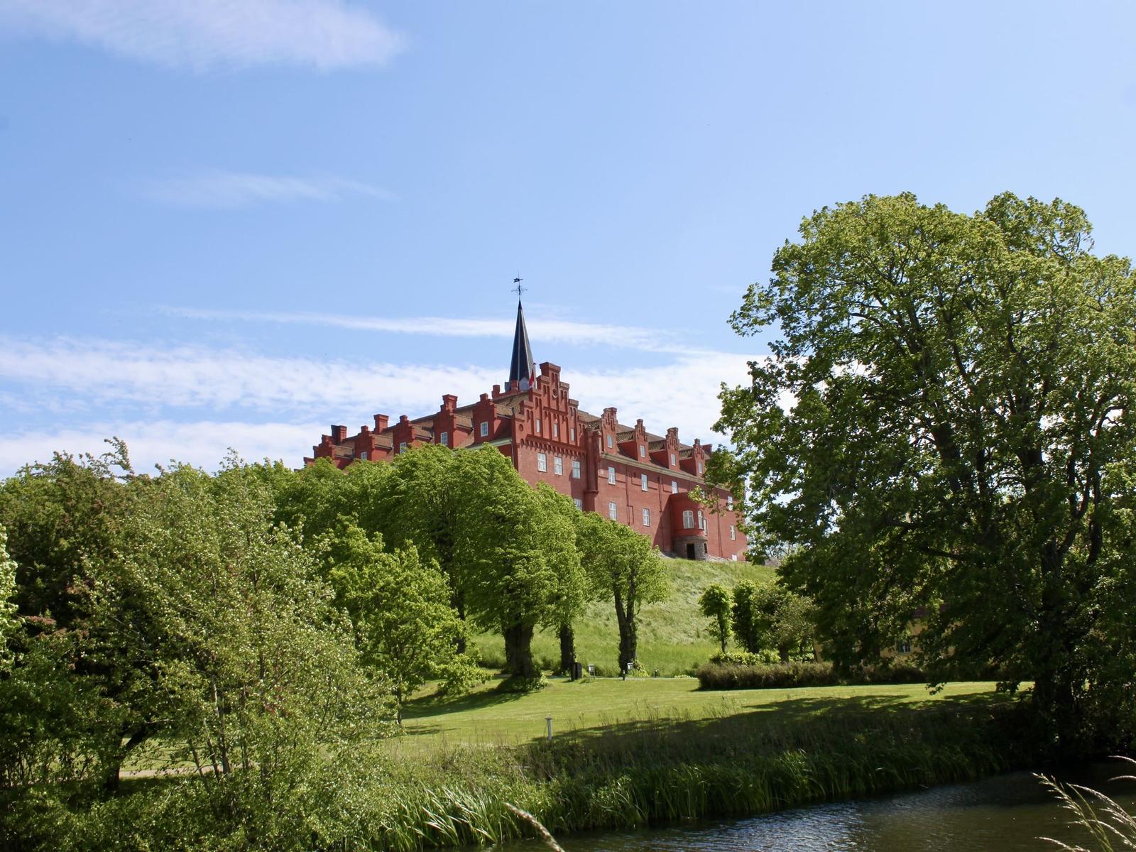 Rotes Schloss auf Hügel mit Grünflächen und Fluss im Vordergrund.