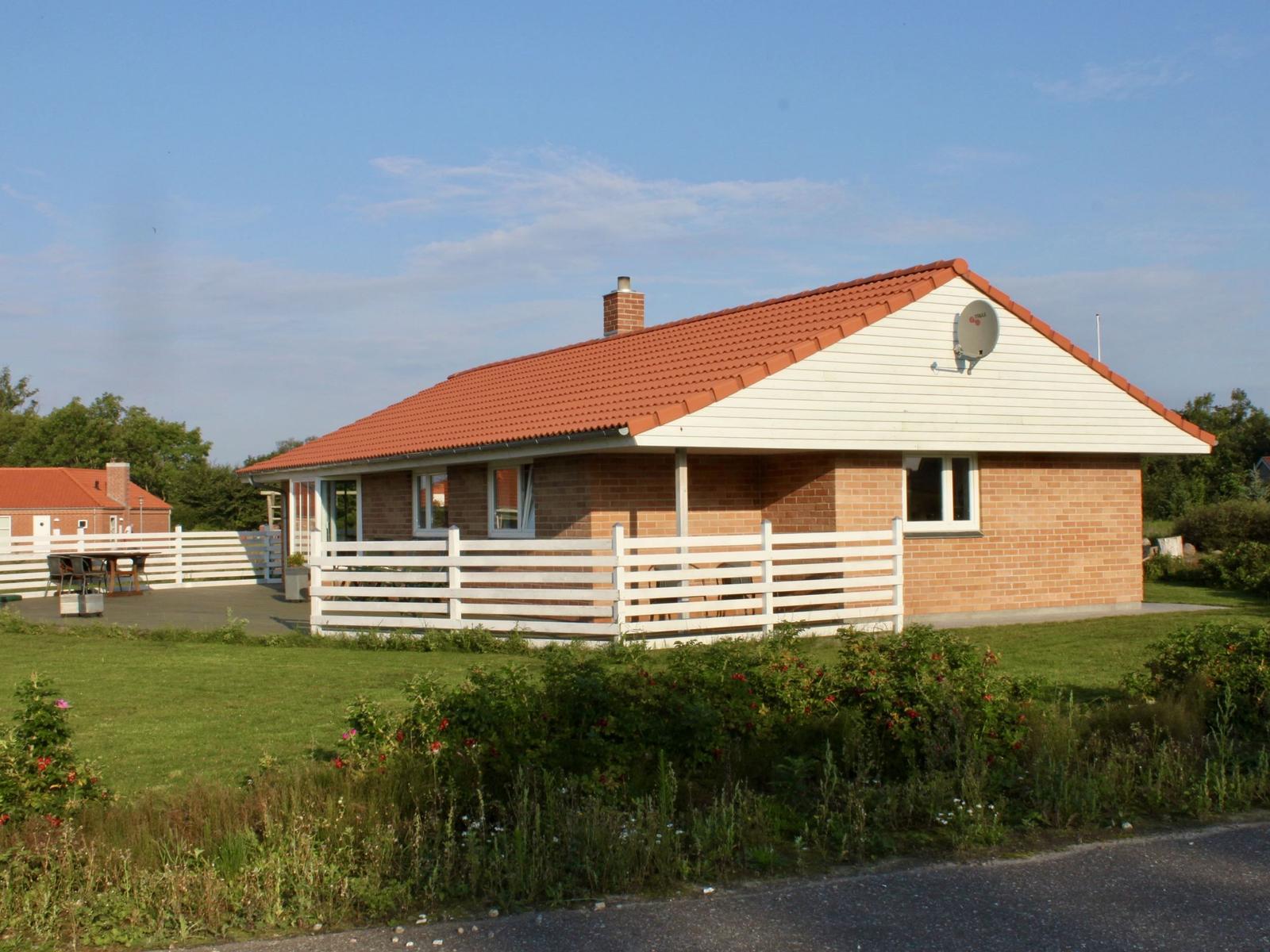 Ein Haus mit rotem Dach, weißer Fassade und weißem Zaun. Grünflächen und ein Satellitenreceiver sind sichtbar.