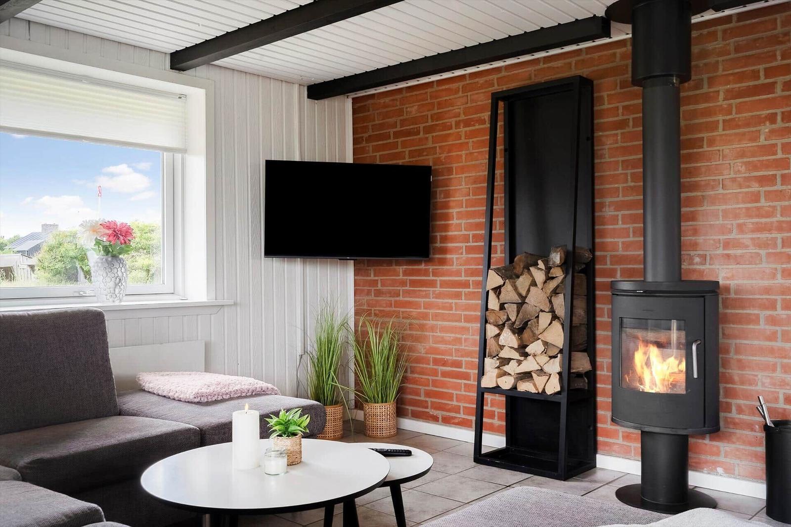 Wohnzimmer mit Kamin, Fernseher und Sitzgelegenheit. Holzstapel und Pflanzen sind sichtbar.