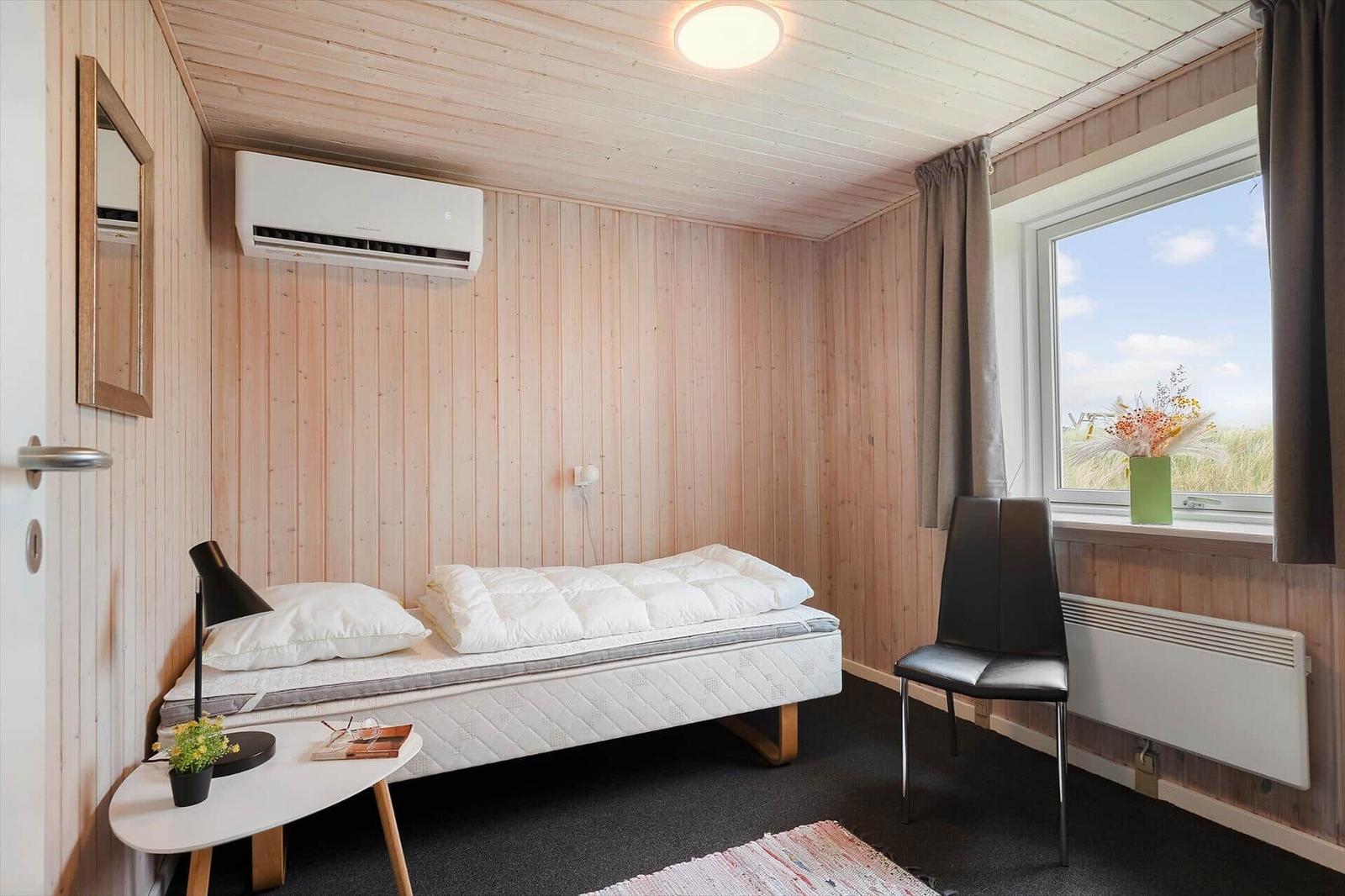 Schlafzimmer mit Bett, Stuhl und Fenster. Holzwände und Heizung sichtbar.