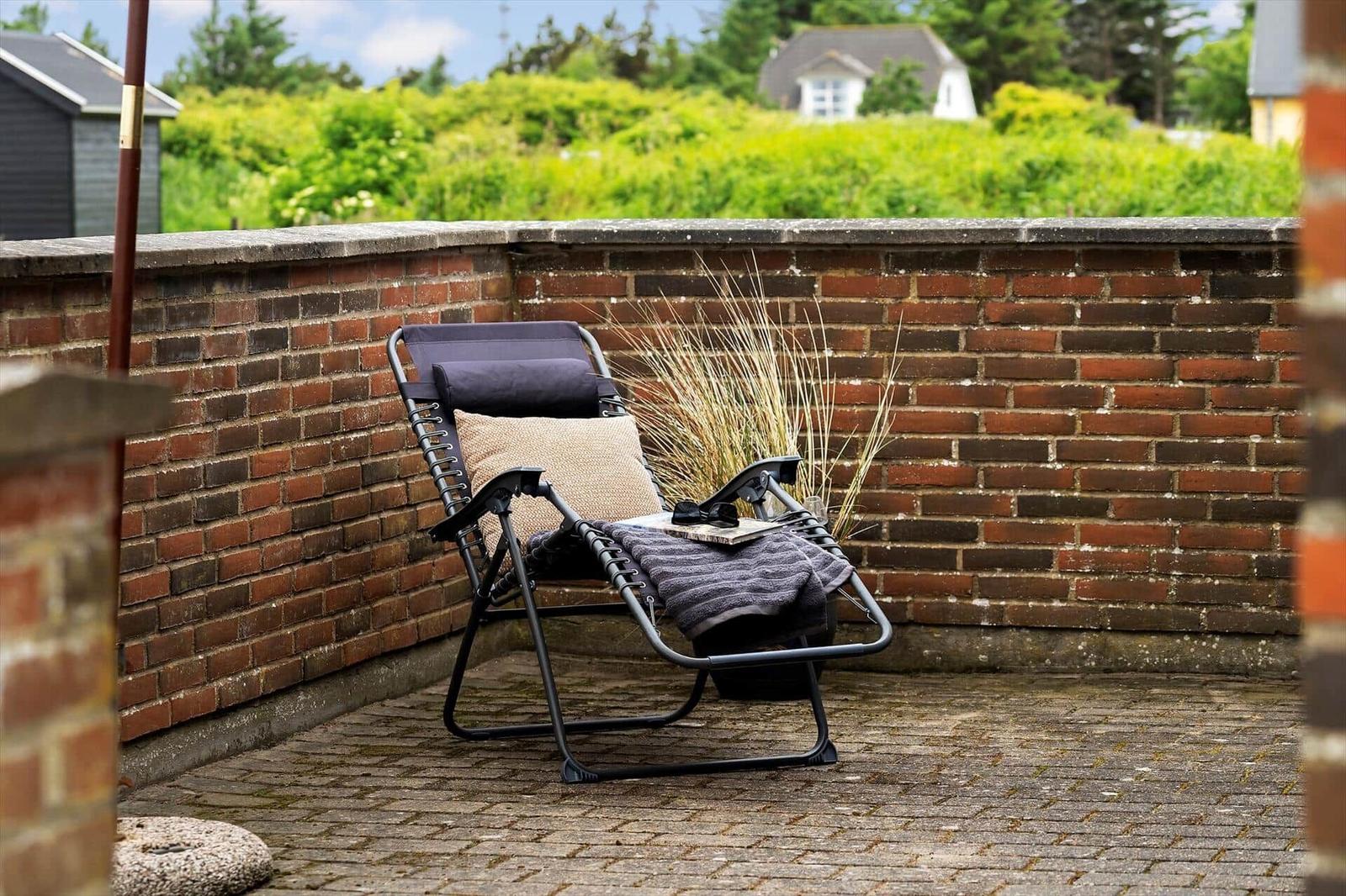 Liegende Sonnenliege auf Terrasse mit Steinmauer und Grünflächen im Hintergrund.