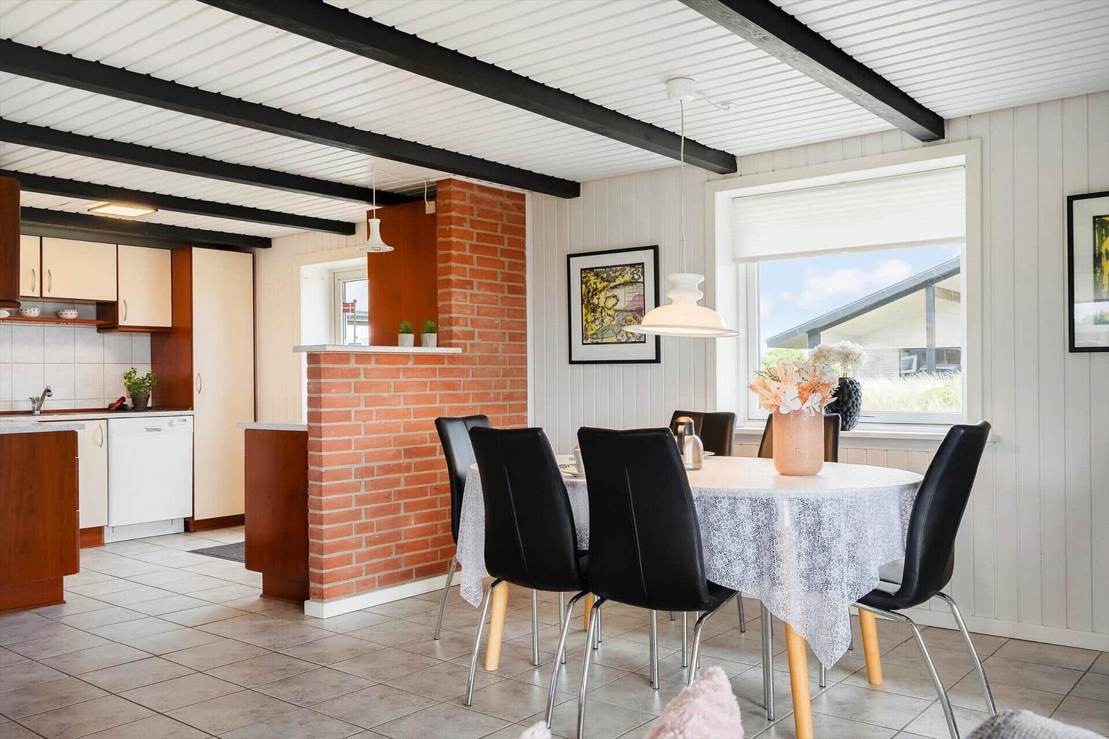 Esszimmer mit runden Tisch, schwarzen Stühlen und Fenster. Küche im Hintergrund. Zentraler Backsteinpfeiler.