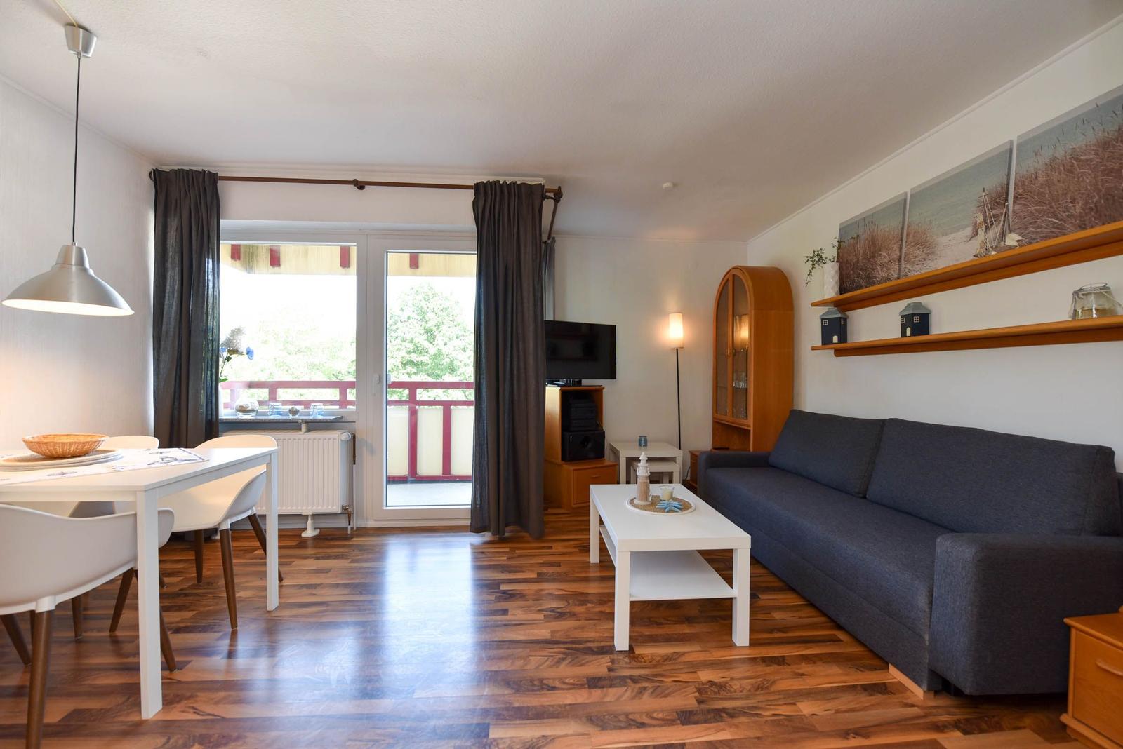 Wohn- und Essbereich mit Sofa, Tisch, TV und Balkon. Holzboden und Wandbilder.
