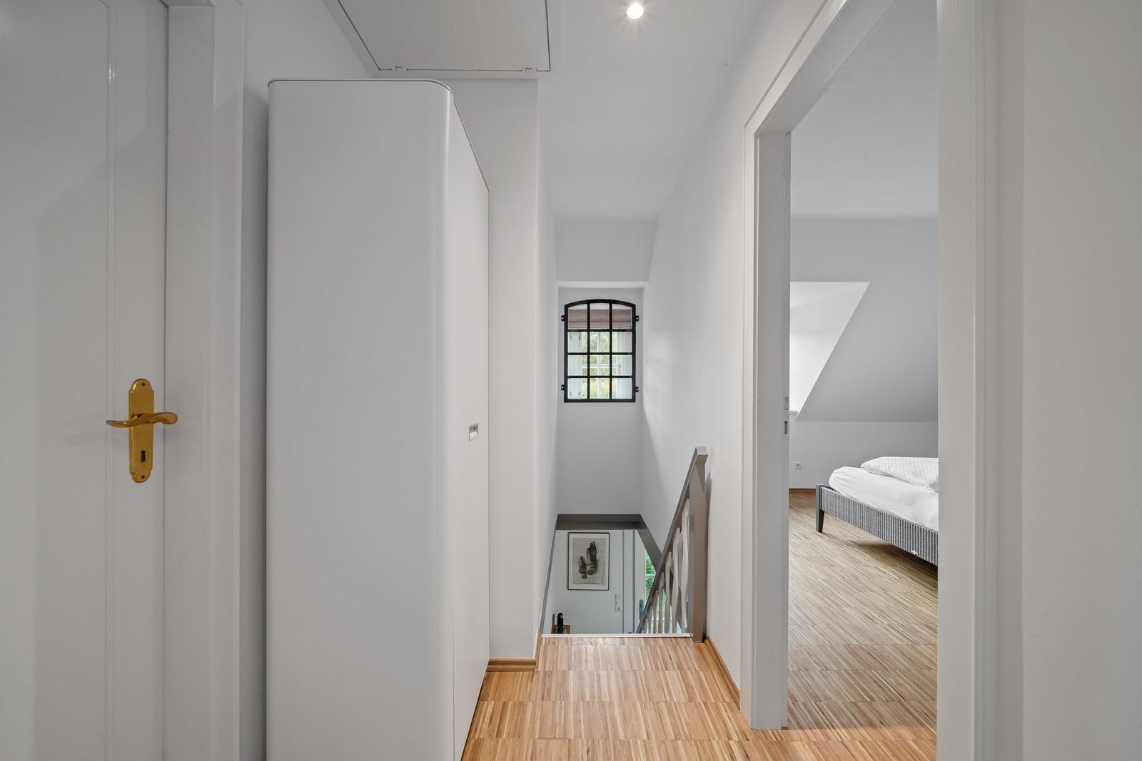 Flur mit Holzboden, Treppenhaus und Blick in Schlafzimmer mit Bett.