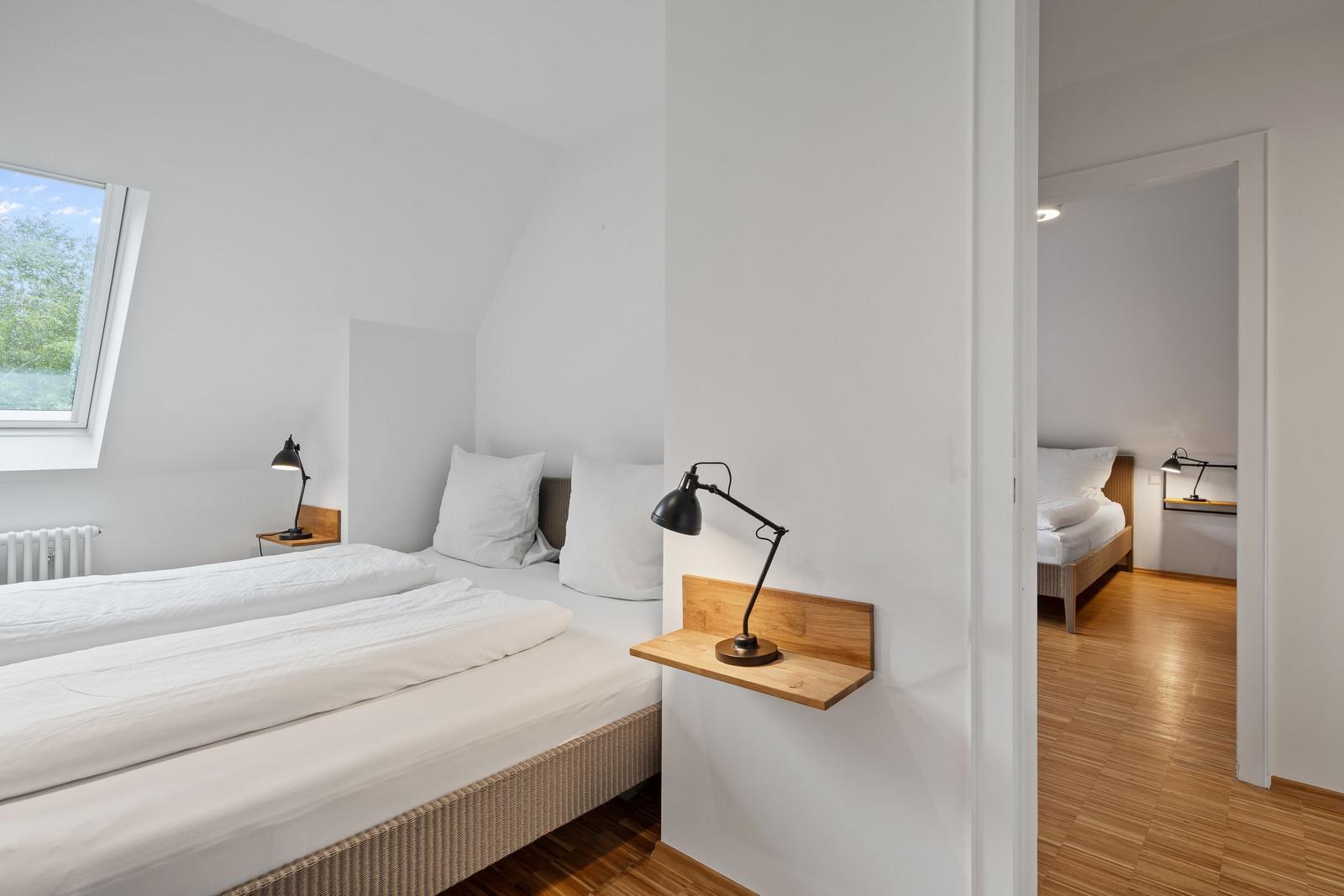 Zimmer mit Dachschrägen, zwei Betten und Holzfußboden. Fenster mit Blick auf Grün.