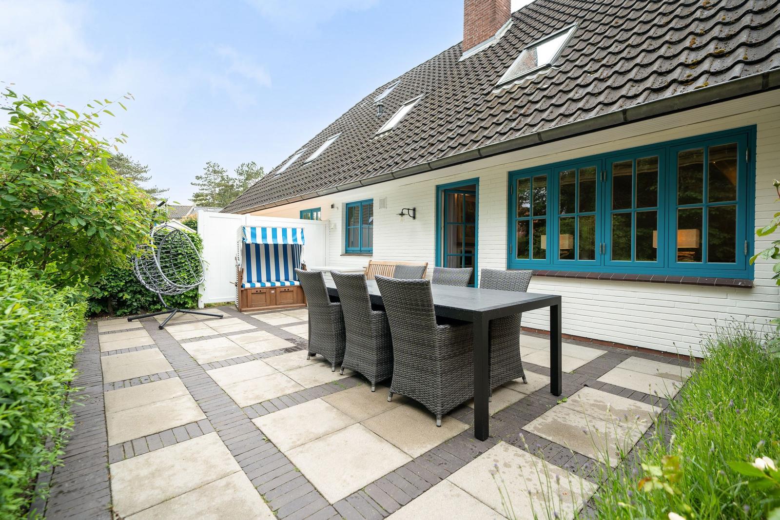 Terrasse mit Tisch, Stühlen und Strandkorb. Haus mit blauen Fenstern und Dachgauben.