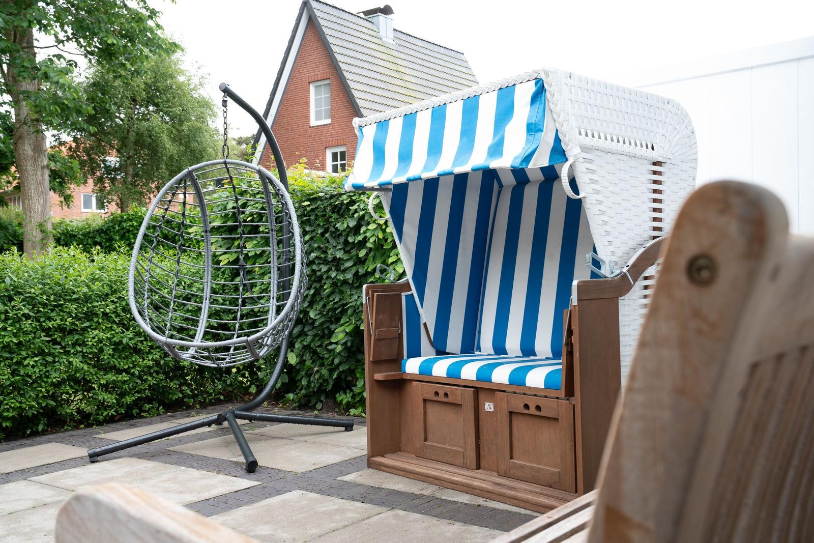 Terrasse mit Strandkorb und Hängesessel. Hintergrund: grüner Heckenzaun und Haus.
