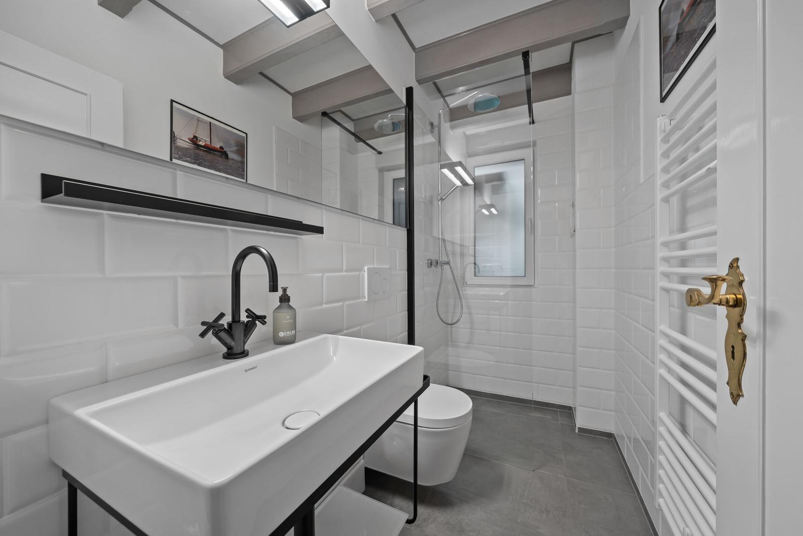 Badezimmer mit Dusche, Waschbecken und Toilette. Weiße Fliesen, schwarze Armatur und goldener Türknauf.