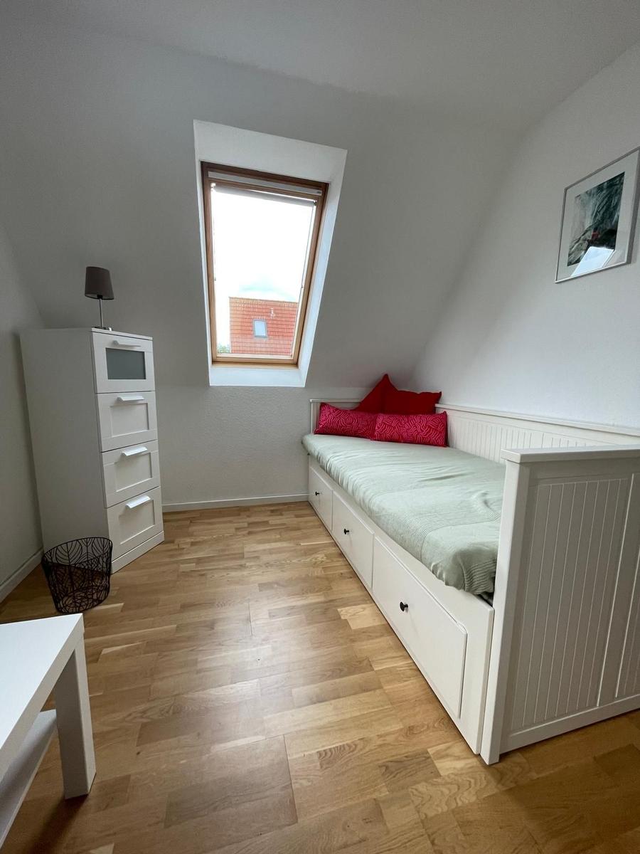 Schlafzimmer mit Dachschräge, weißem Bett und Holzboden. Fenster und Schrank sind sichtbar.