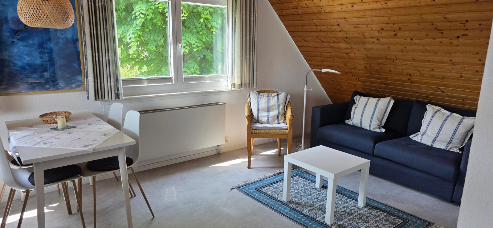 Wohn- und Essbereich mit Sofa, Tisch und Stühlen. Holzwand und Fenster mit Blick auf Grün.