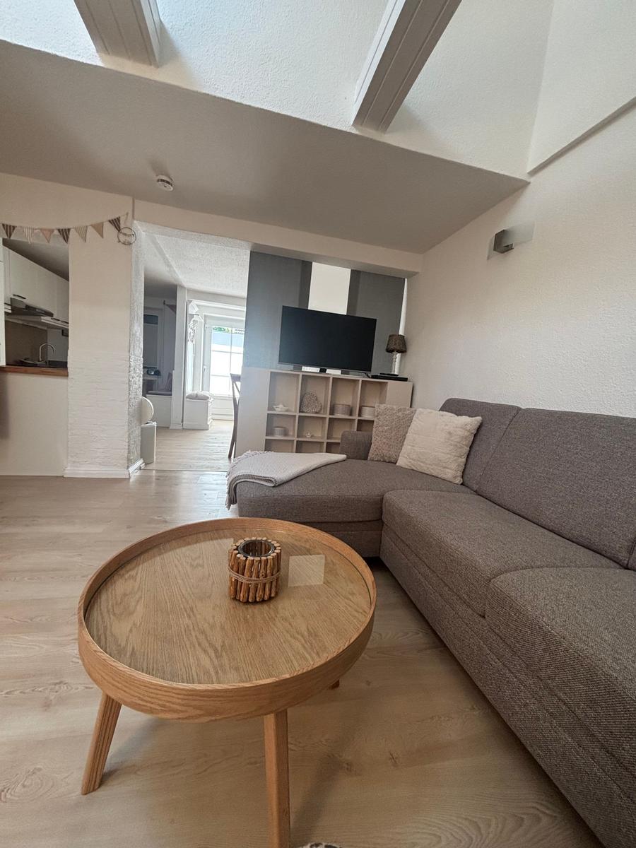 Wohnbereich mit grauem Sofa, runden Tisch und Fernseher. Küche im Hintergrund.