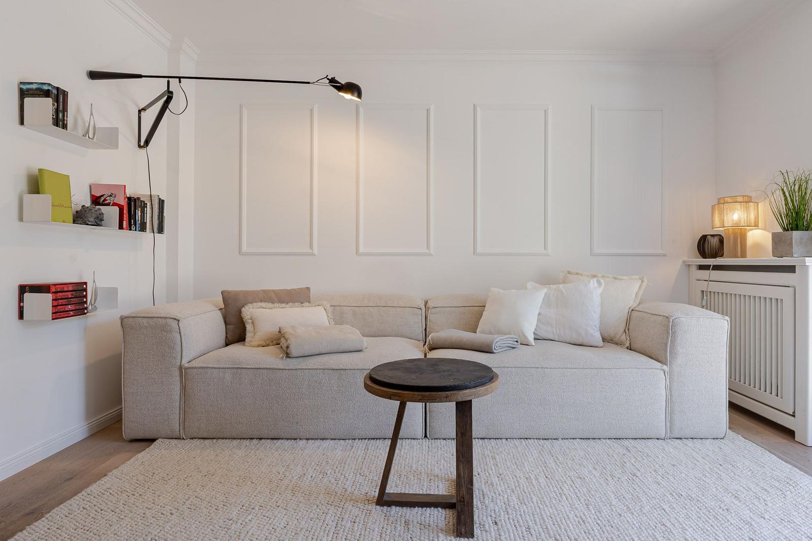 Ein Wohnzimmer mit einem hellen Sofa, einem runden Tisch und einer Wandlampe.