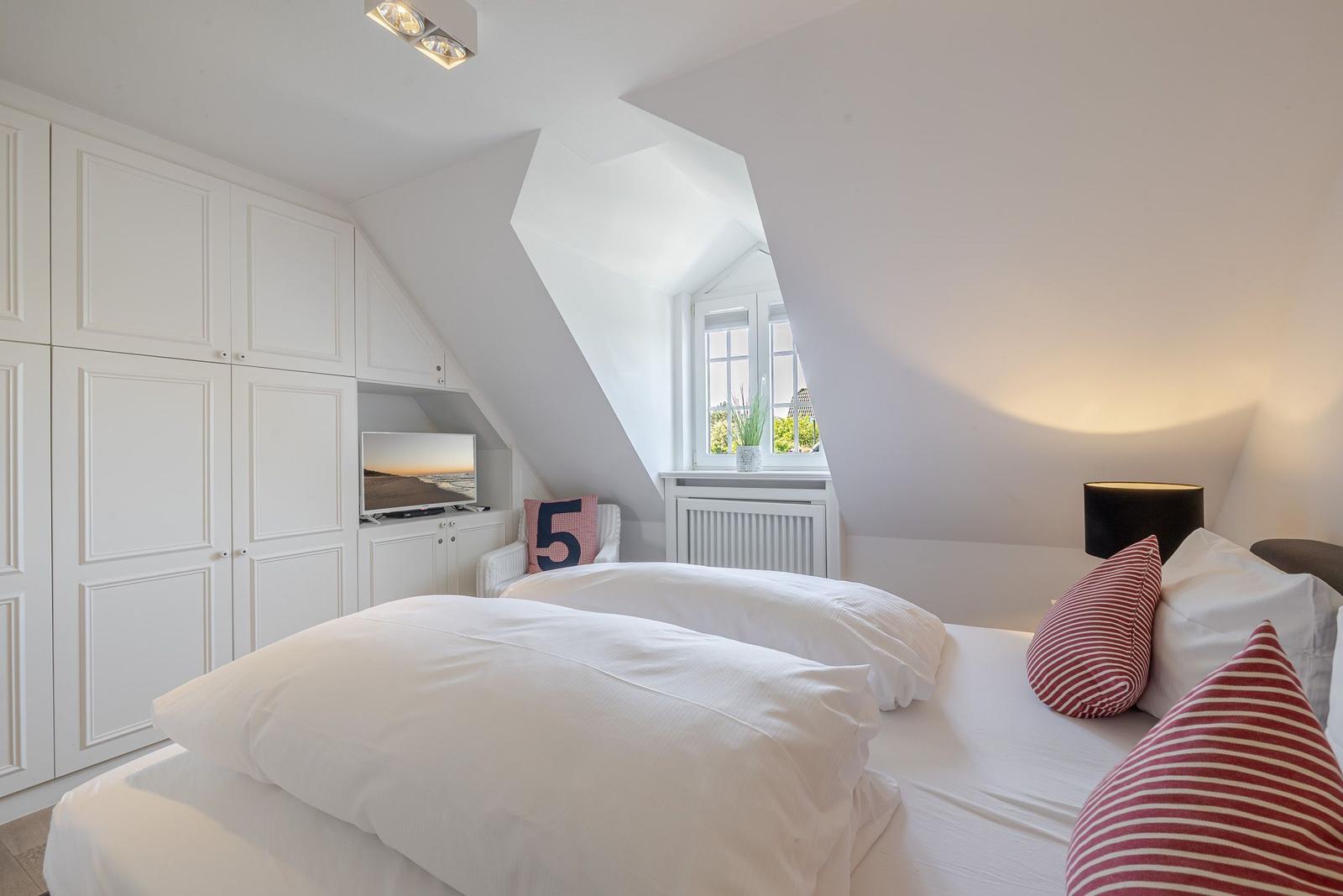 Schlafzimmer mit Dachschrägen, weißem Bett und Kleiderschrank. Fenster mit Pflanze und Fernseher.