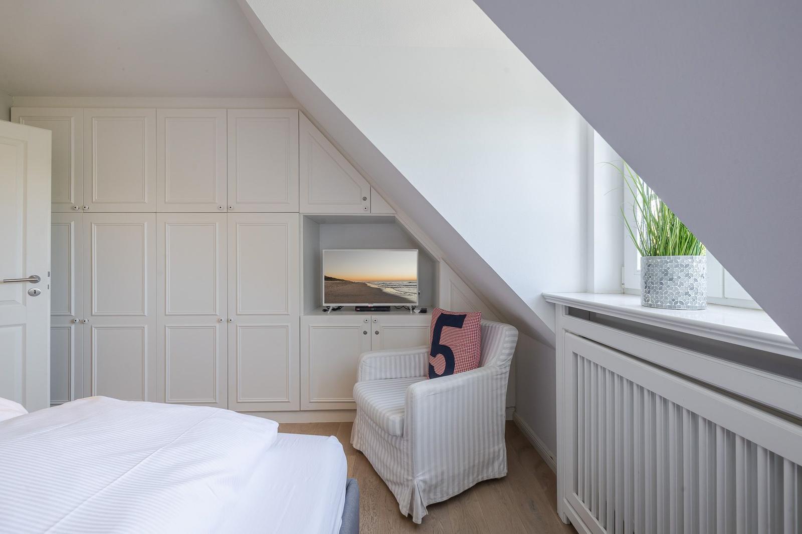 Schlafzimmer mit Dachschräge, weißer Einbauschränke, Sessel und Fernseher.