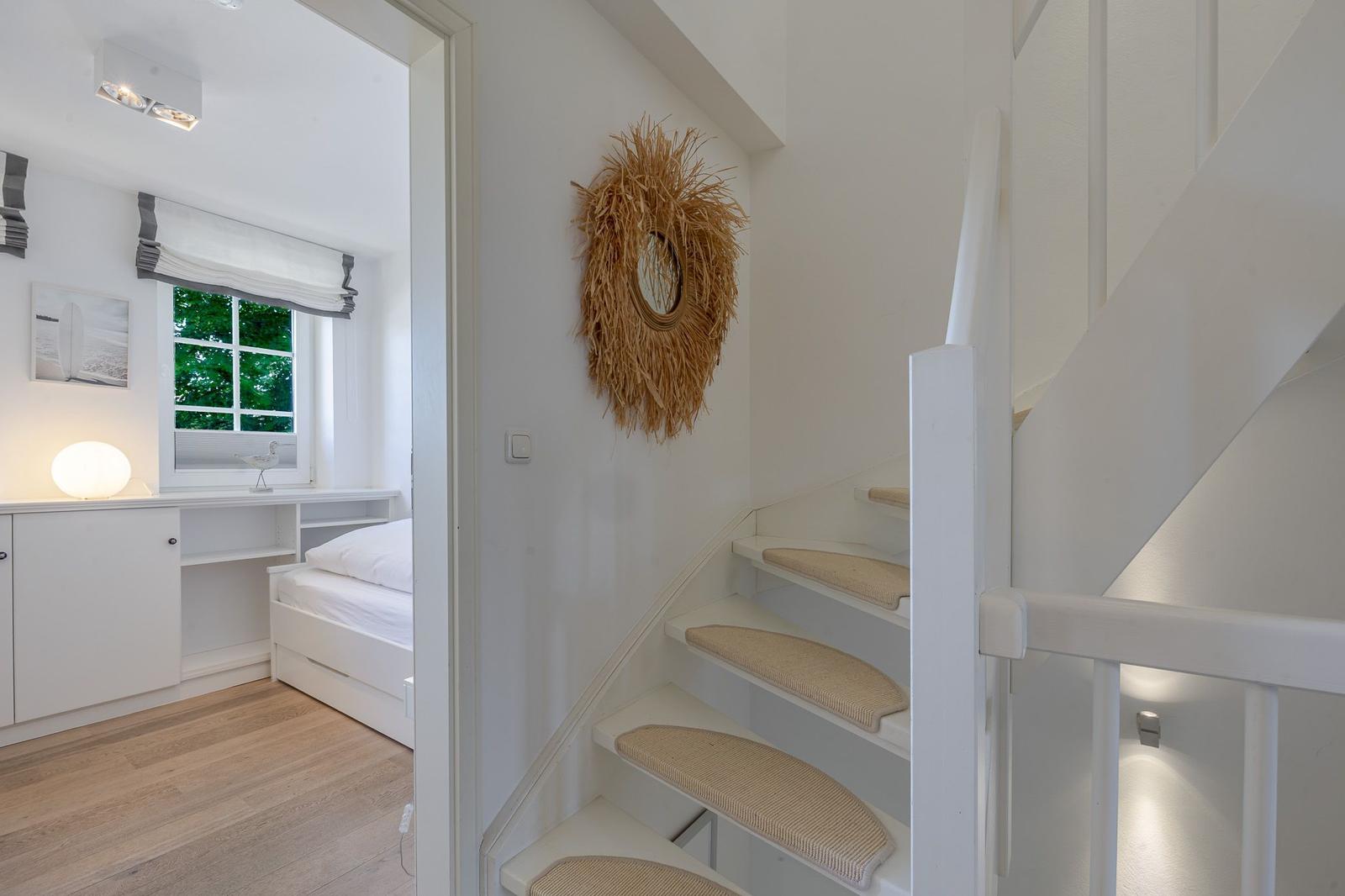 Treppenhaus mit weißer Treppe und Sitzmatte. Im Hintergrund ist ein Schlafzimmer mit Fenster und Schrank.