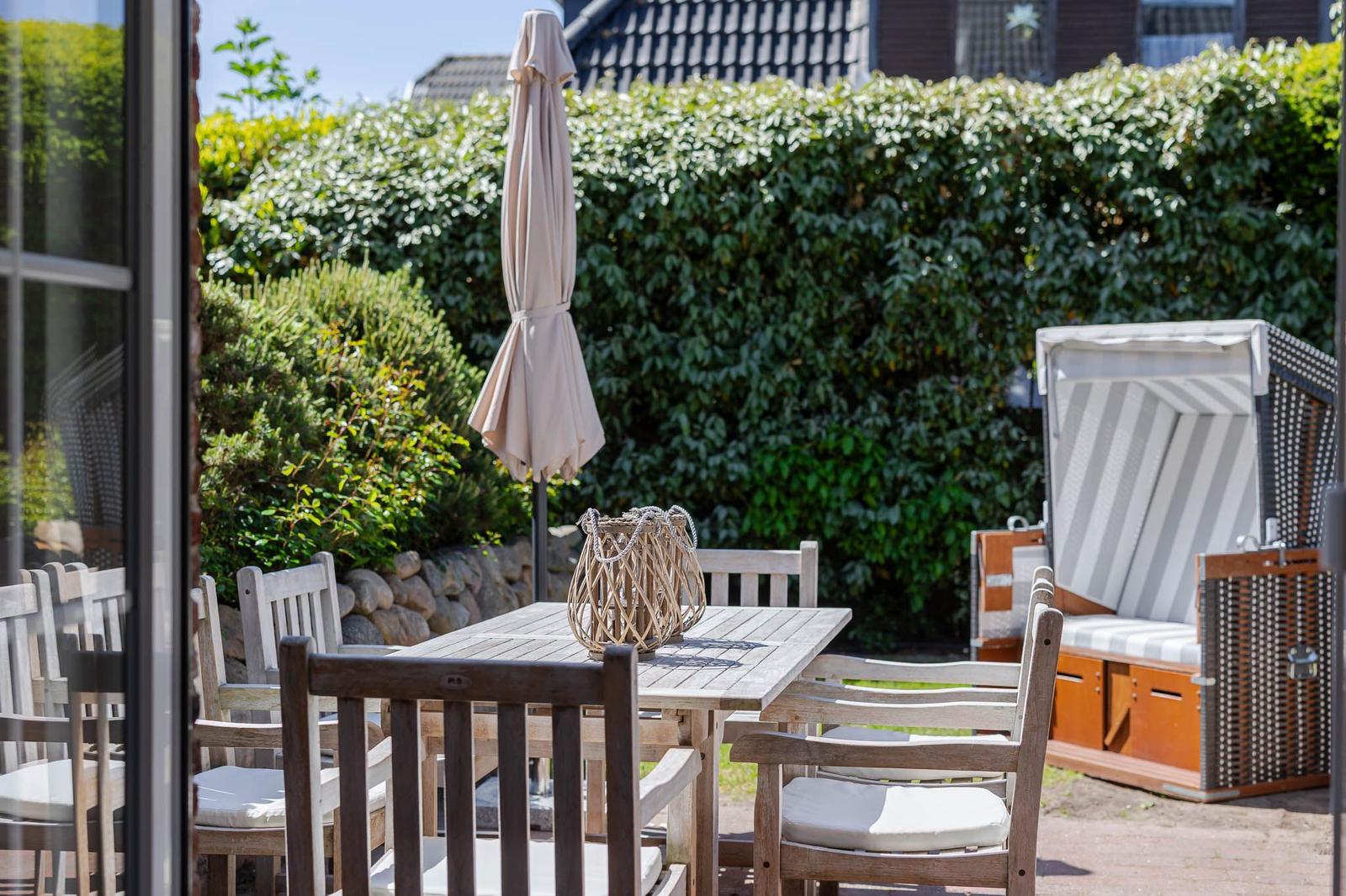 Terrasse mit Holztisch, Stühlen und geschlossenem Sonnenschirm. Im Hintergrund grüne Hecke und Strandkorb.