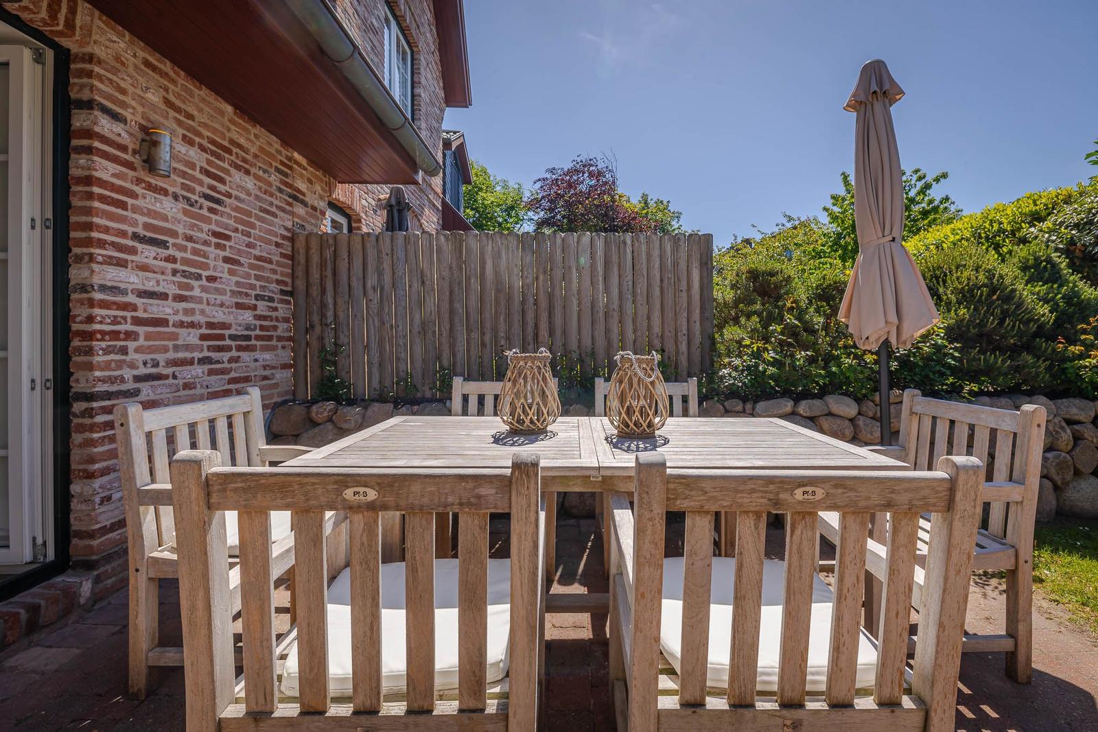 Holztisch mit sechs Stühlen und weißem Sitzkissen auf Terrasse neben Backsteinhaus.