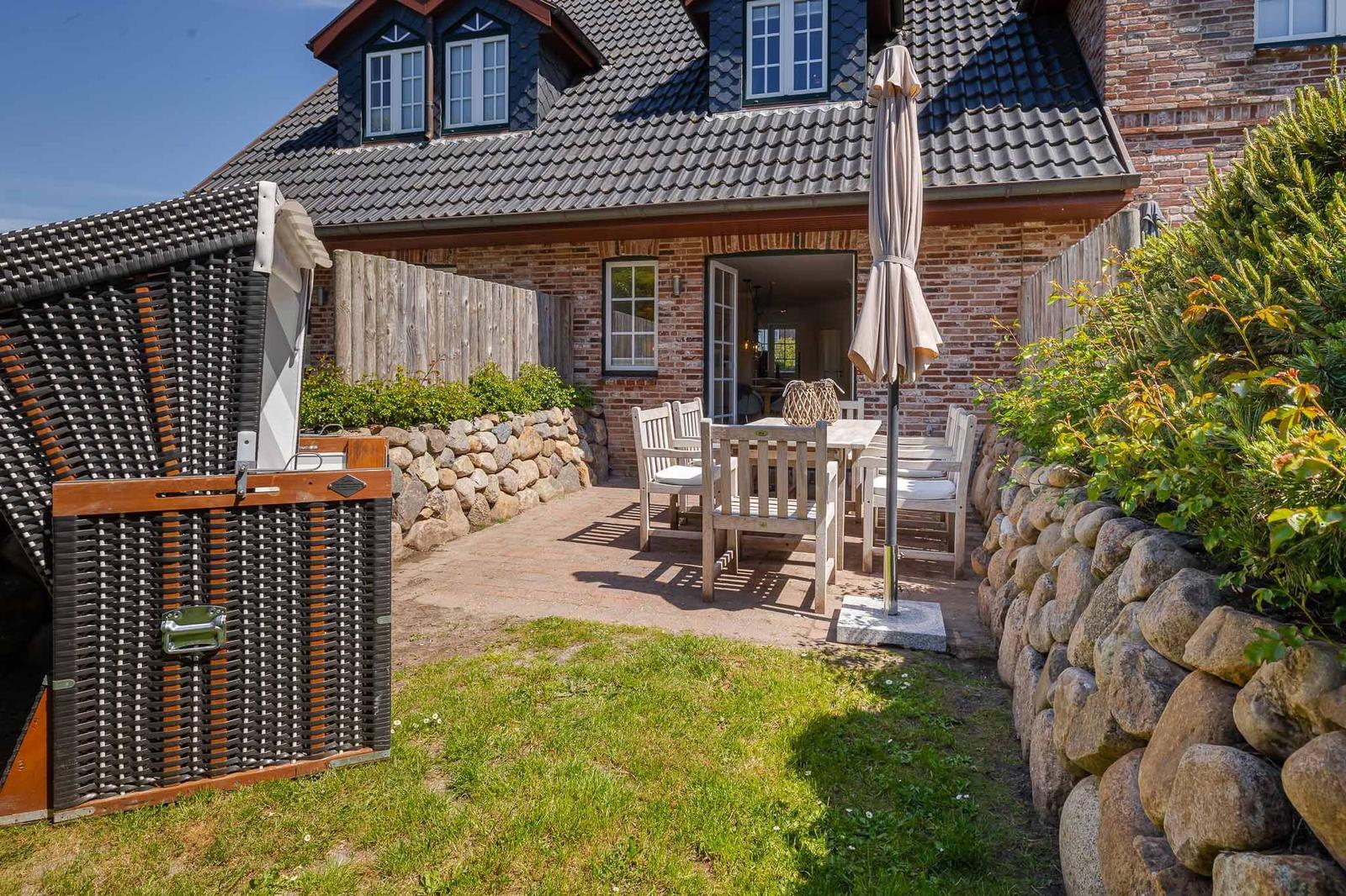 Haus mit Terrasse, Garten und Holzgartenmöbeln. Steinmauer und Holzzaun umgeben den Außenbereich.
