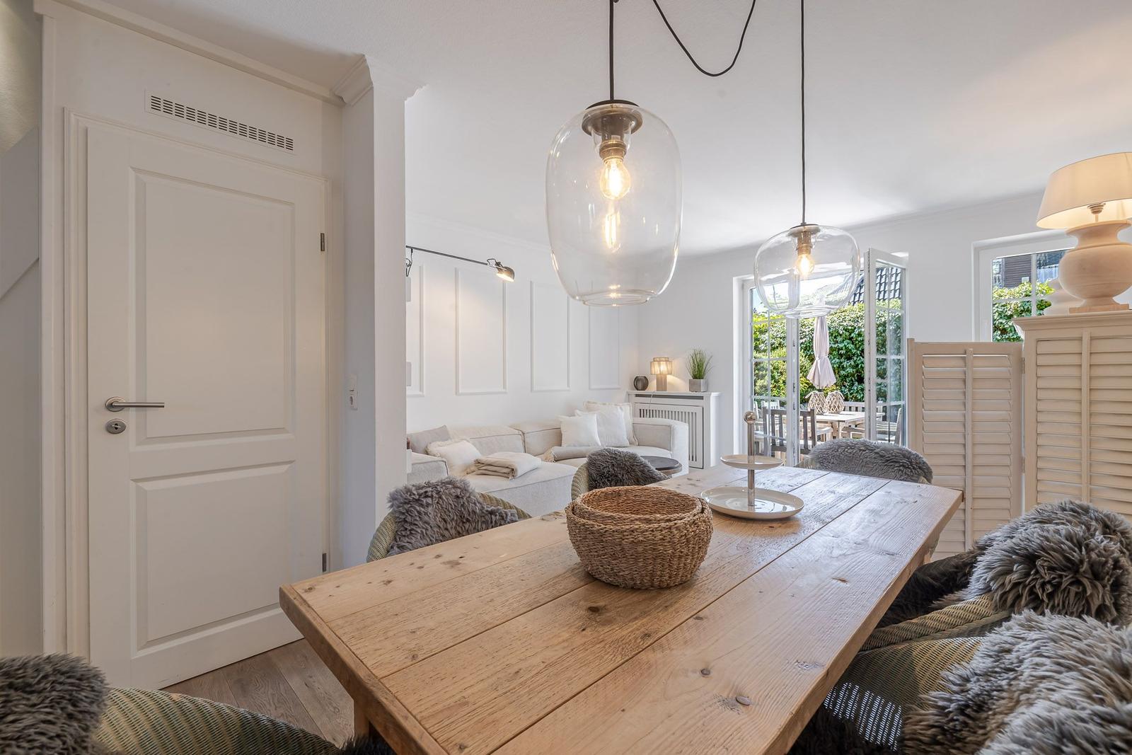 Essbereich mit Holztisch und hängenden Lampen. Durchgang zur Wohnfläche mit Sofa und Fenster.