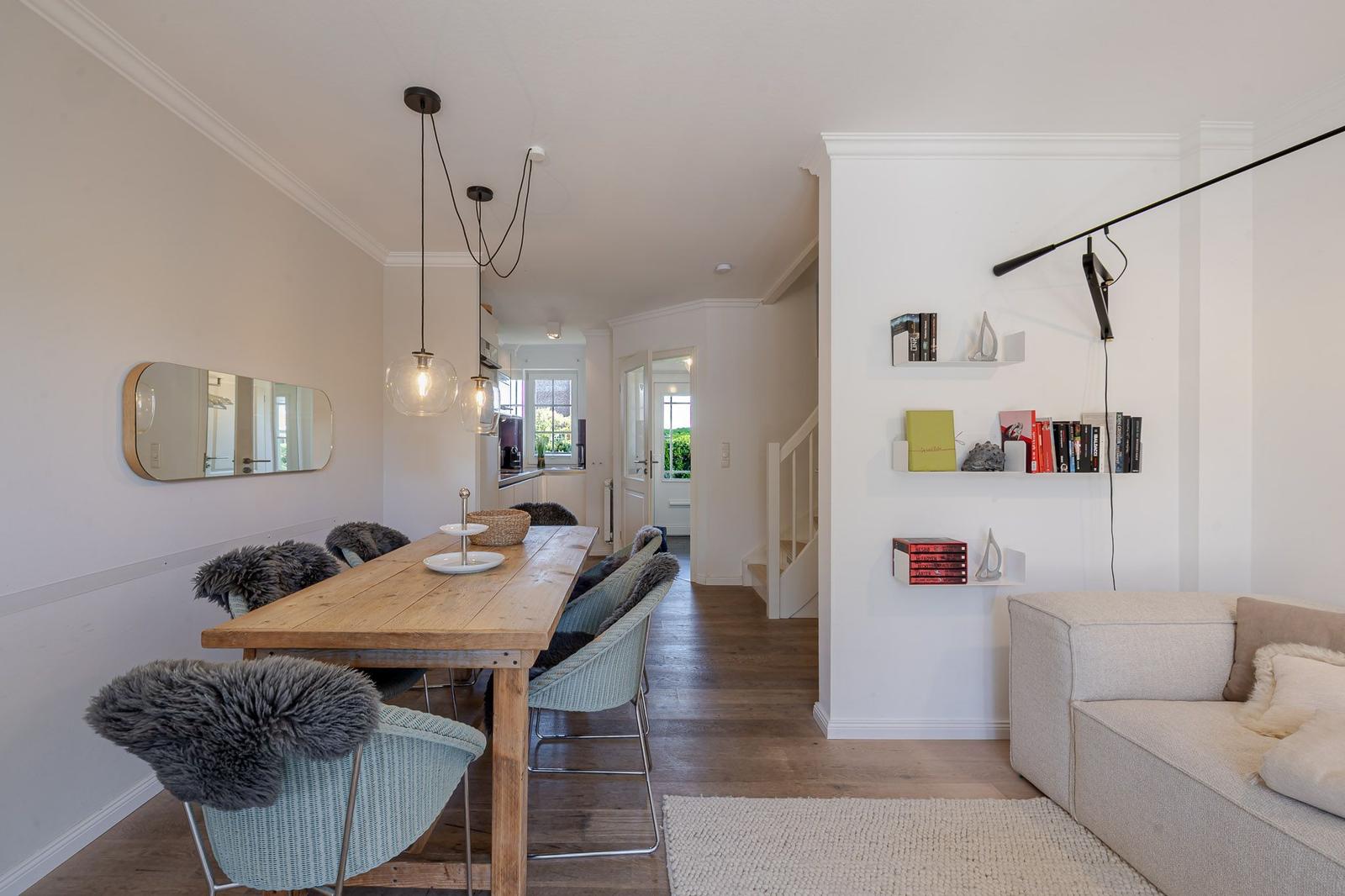Essbereich mit Holztisch und Stühlen, Wohnbereich mit Sofa und Regal. Helle Wand und Holzboden.