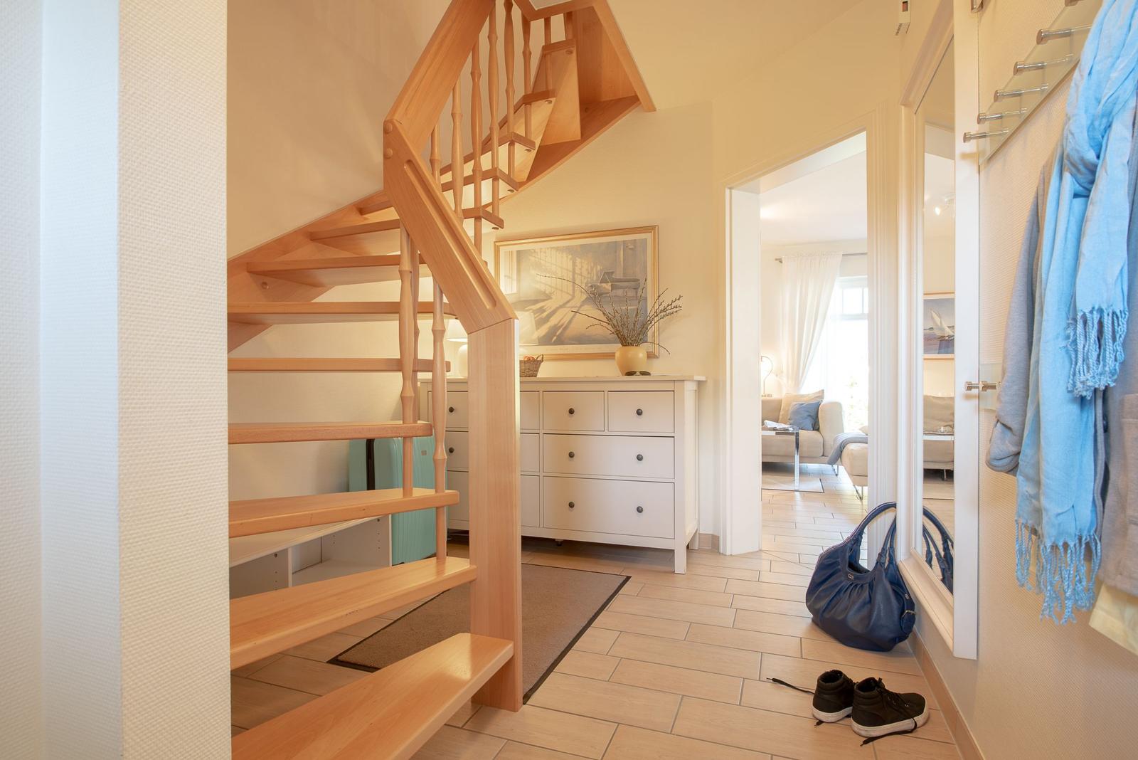 Hausflur mit Holztreppe, Schuhen und Kleidung. Im Hintergrund Wohnzimmer.