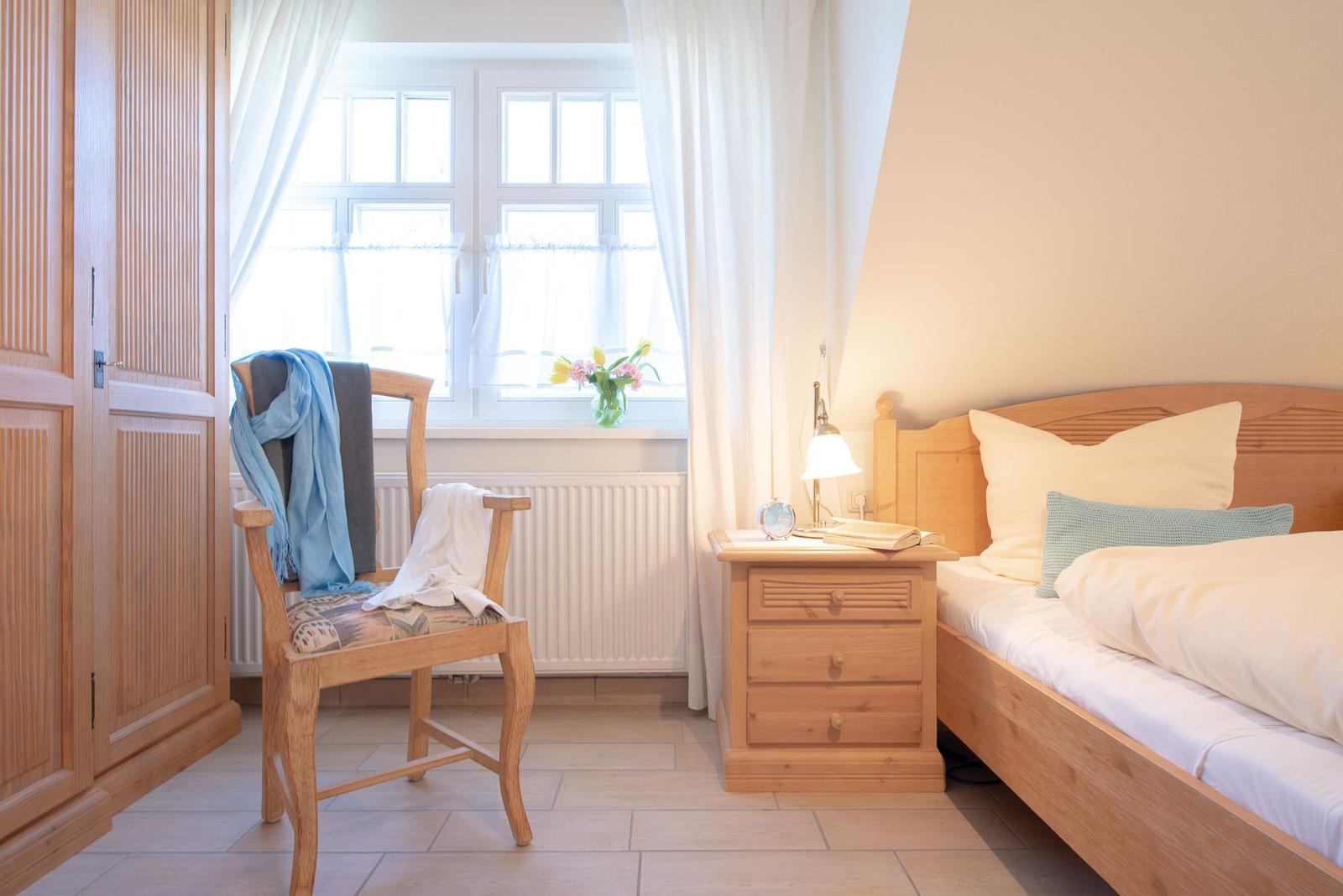 Ein Schlafzimmer mit Holzbett, Nachttisch und Stuhl. Fenster mit Vorhängen und Blumen auf dem Fensterbrett.