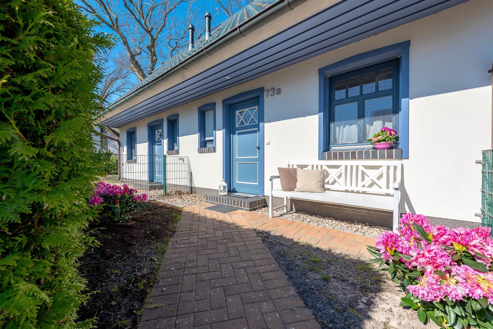 Haus mit blauen Fenstern und Tür, weißer Bank mit Kissen, Blumenbeete und gepflastertem Weg.