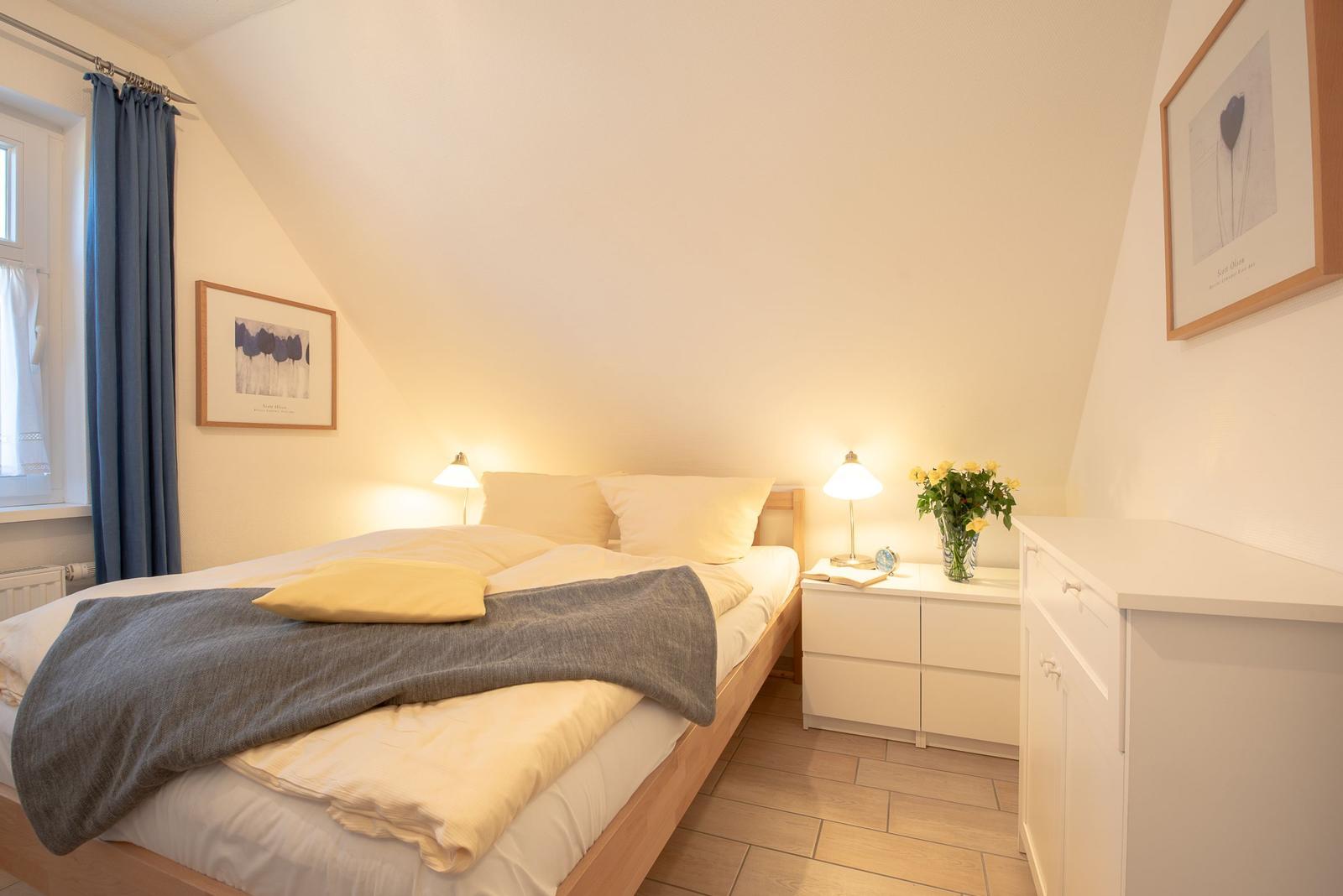 Schlafzimmer mit Dachschrägen, Bett, Nachttisch und Fenster mit Vorhängen.