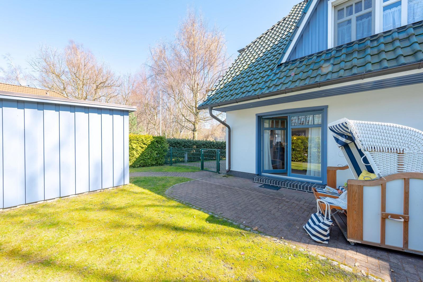Haus mit Terrasse, Garten und Liegestühlen. Blaue Fensterrahmen und grüner Dachfirst.