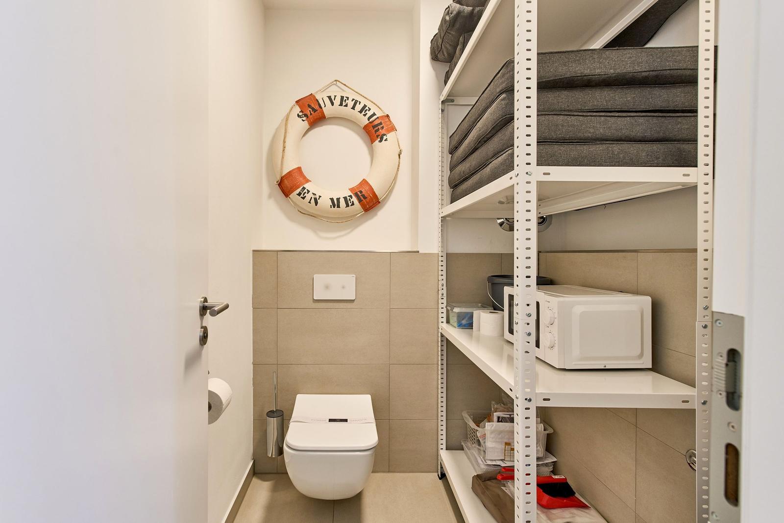 Toilette mit Rettungsring an der Wand und Metallregal mit Kissen und Mikrowelle.