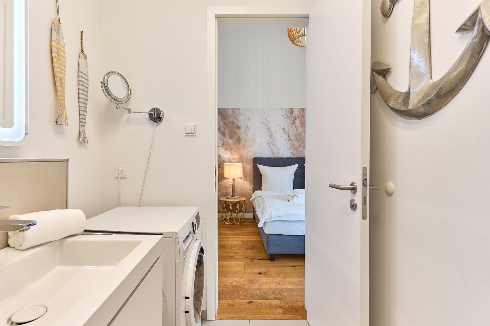 Badezimmer mit Waschmaschine und Blick in Schlafzimmer mit Bett und Holzboden.