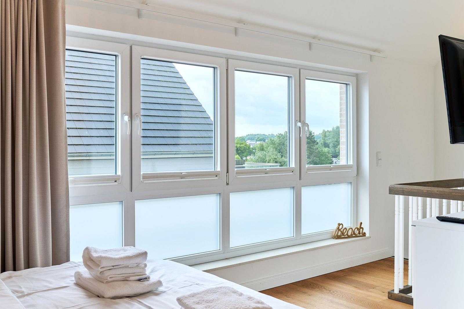 Ein Schlafzimmer mit großem Fenster, Blick auf Dach und Grün, Badetuch auf Bett, Wand-TV.