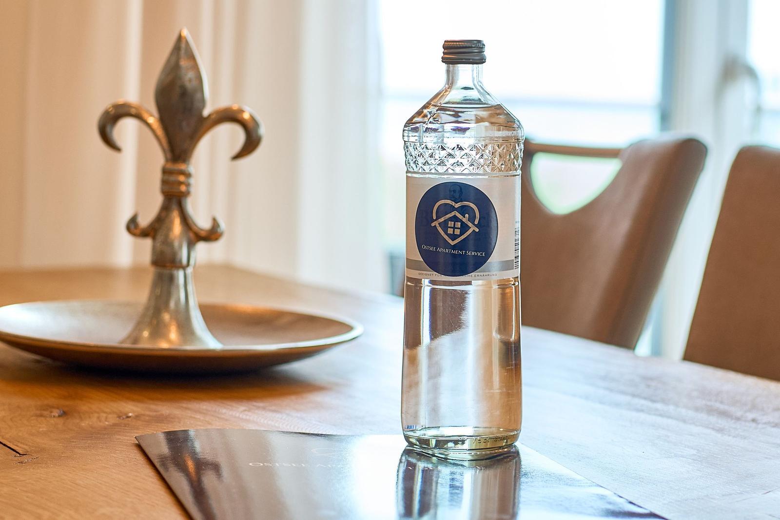 Eine Wasserflasche mit Logo steht auf einem Holztisch neben einem silbernen Fleur-de-Lis-Objekt.