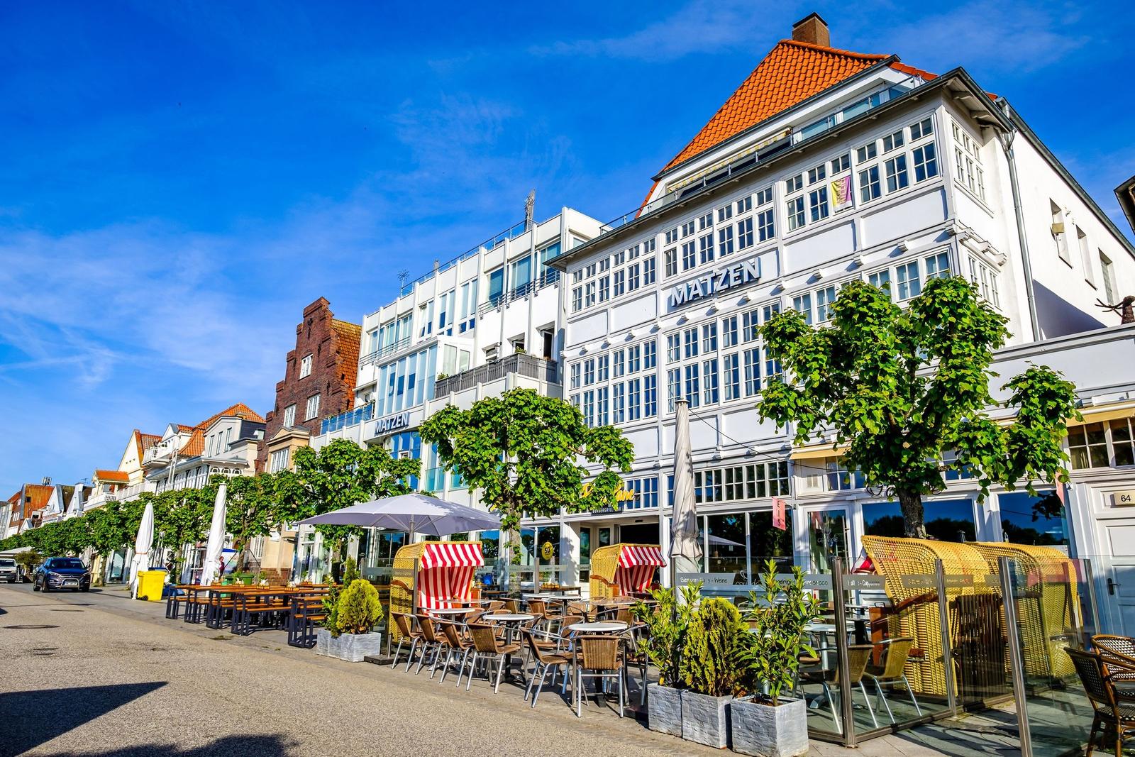 Strandpromenade mit Café, Stühlen und Bäumen unter blauem Himmel.