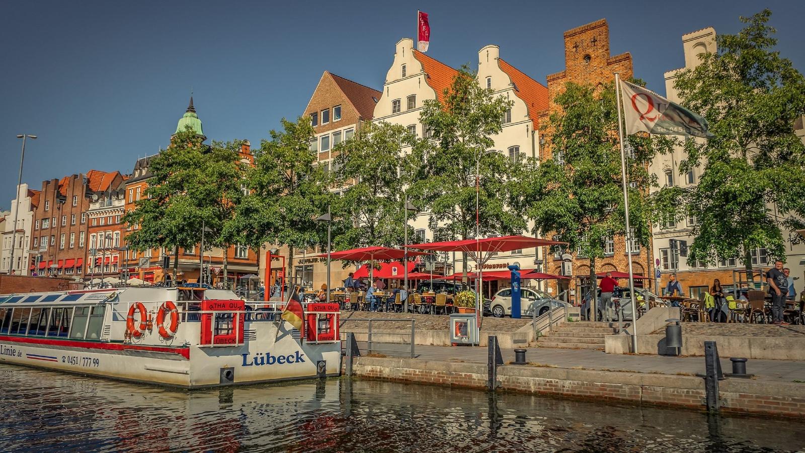 Schiff am Hafen von Lübeck mit historischen Gebäuden und Terrassen im Hintergrund.
