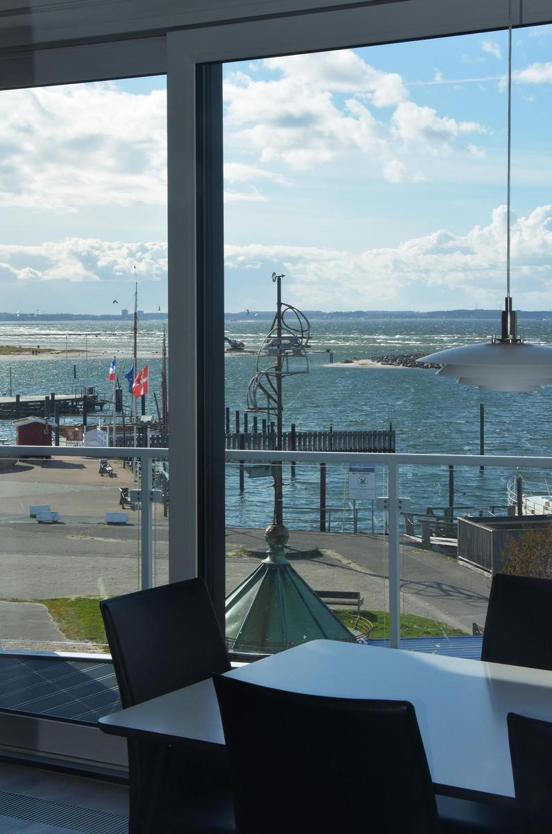 Tisch und Stühle vor großem Fenster mit Blick auf Hafen und Meer.