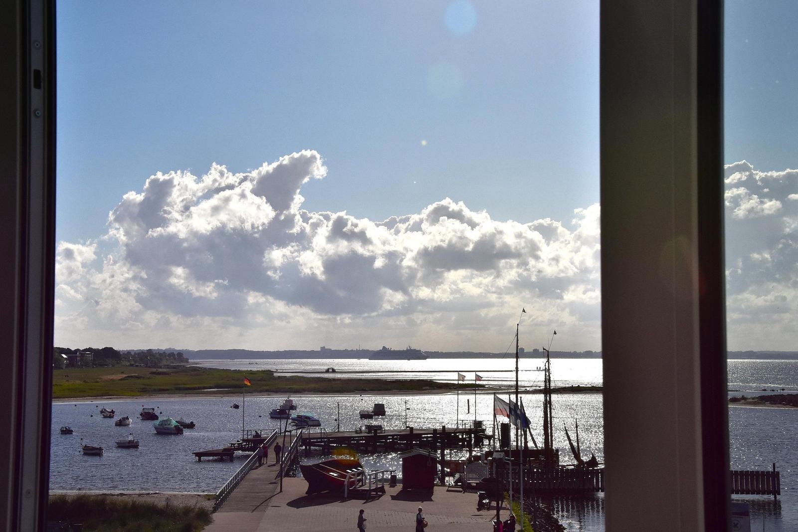 Aus dem Fenster blickt man auf einen Hafen mit Booten und einem Schiffssteg unter blauem Himmel mit Wolken.