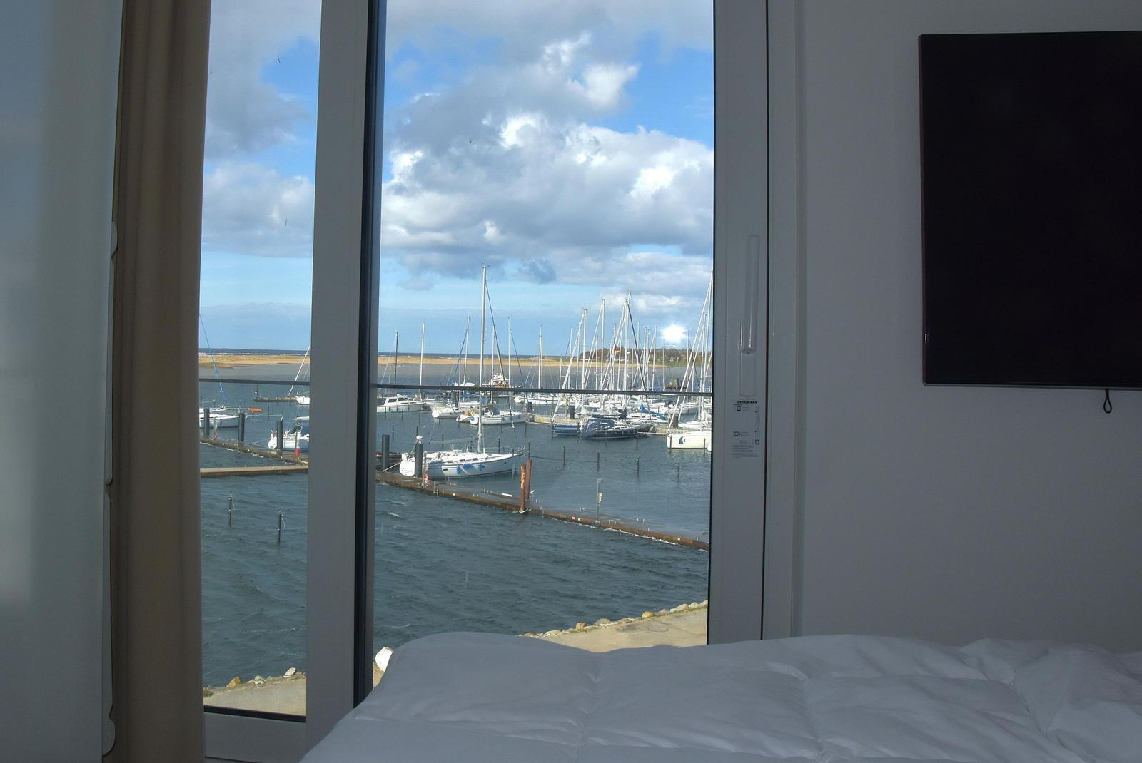 Blick auf Hafen mit Segelbooten durch großes Fenster. Bett und Wandfernseher sichtbar.