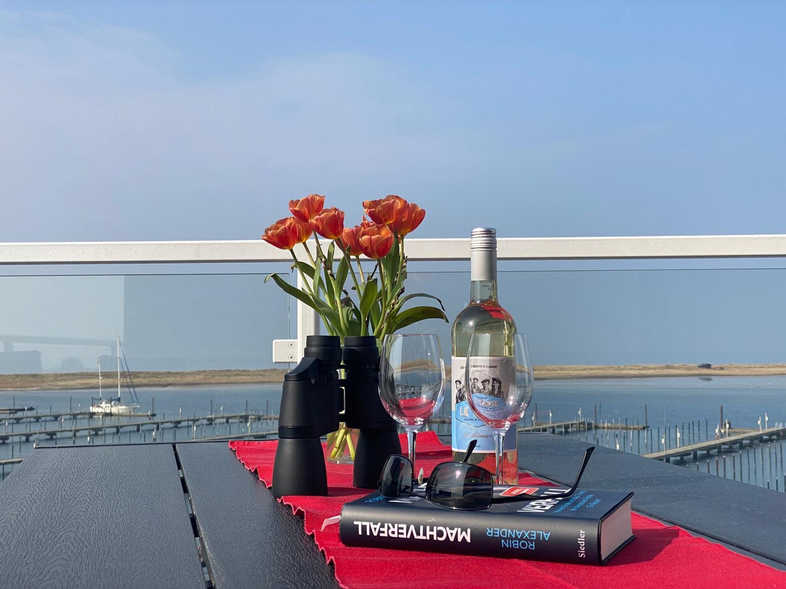 Tisch auf Balkon mit Tulpen, Wein, Fernglas und Buch vor Wasser.