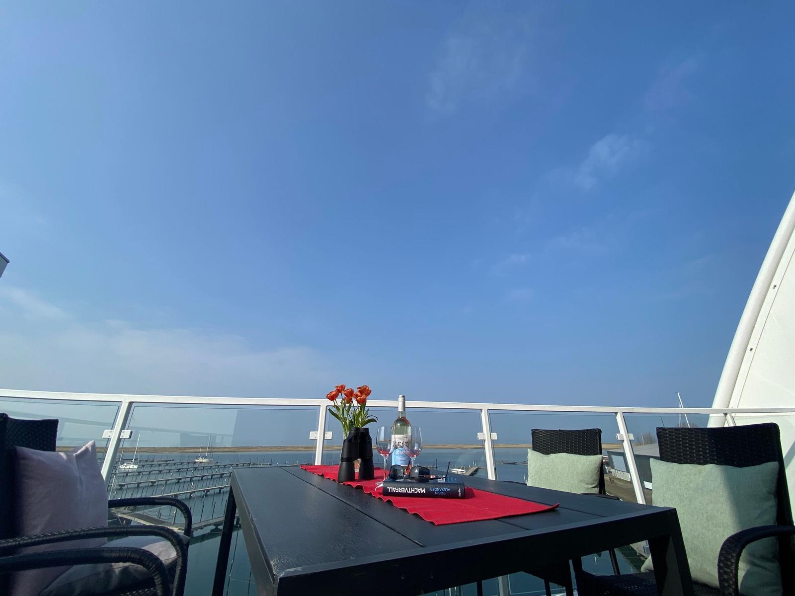 Terrasse mit Tisch, Stühlen und Blick auf Hafen und Himmel.