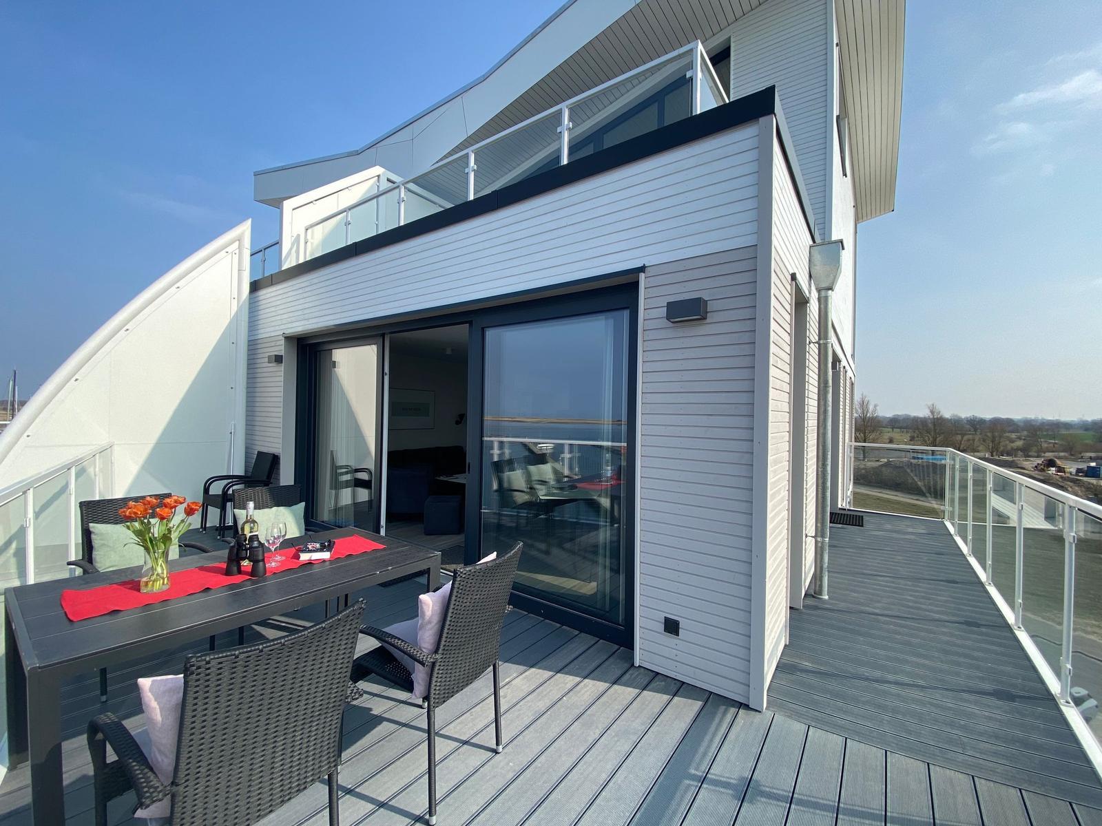 Terrasse mit Tisch, Stühlen und Blumen. Glasfront öffnet sich ins Innere.