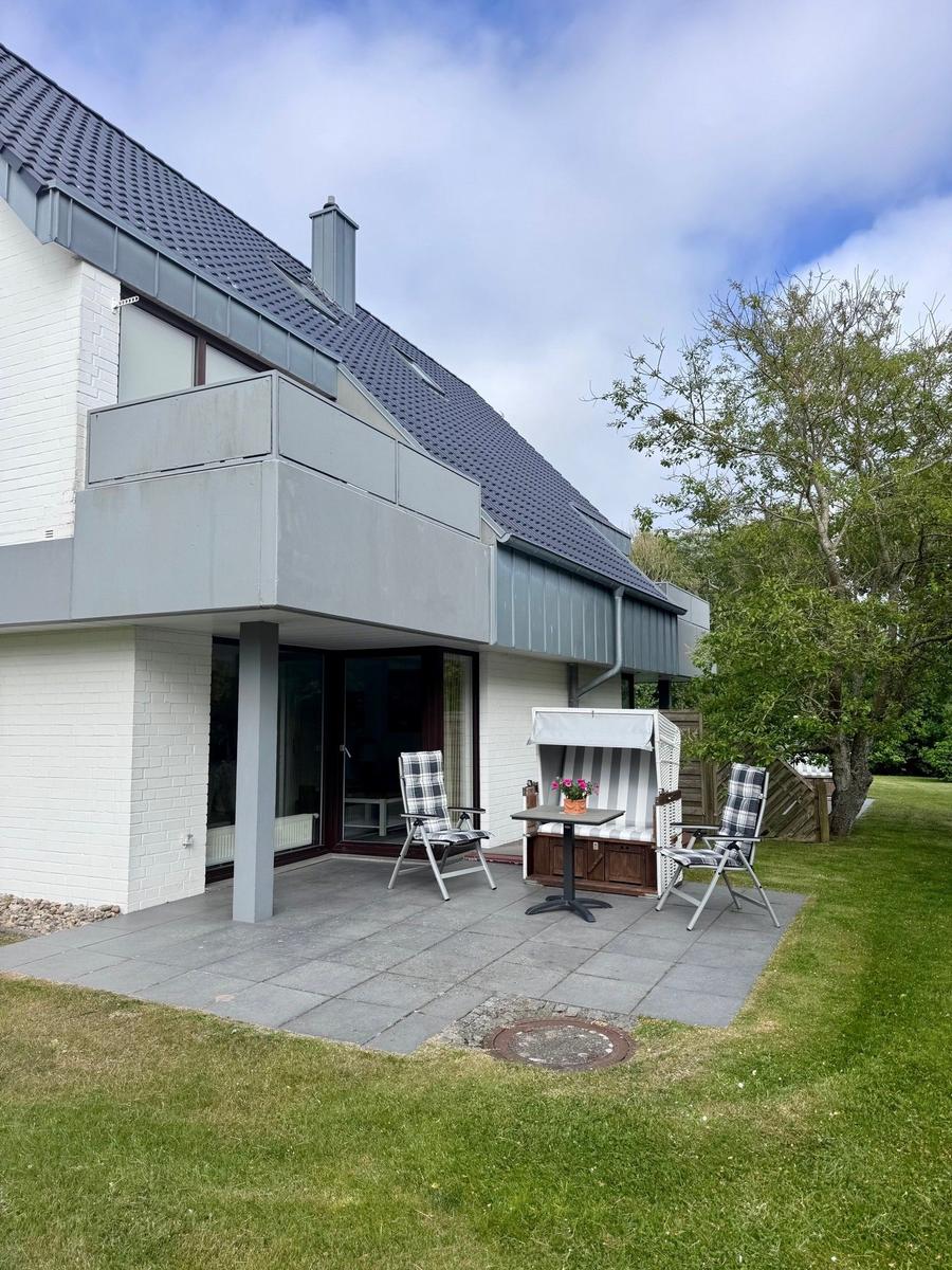 Terrasse mit Stuhl, Tisch und Strandkorb. Hintergrund: Haus mit Balkon und Grünfläche.