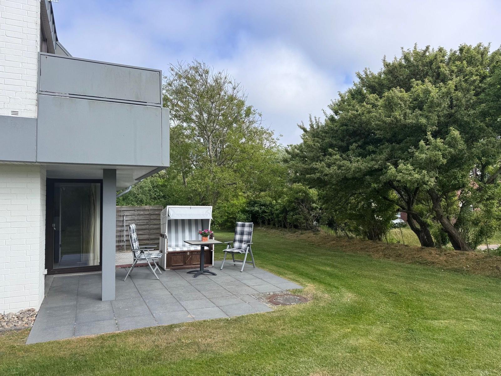 Terrasse mit Gartenhaus, Tisch und zwei Stühlen. Grünfläche und Bäume im Hintergrund.
