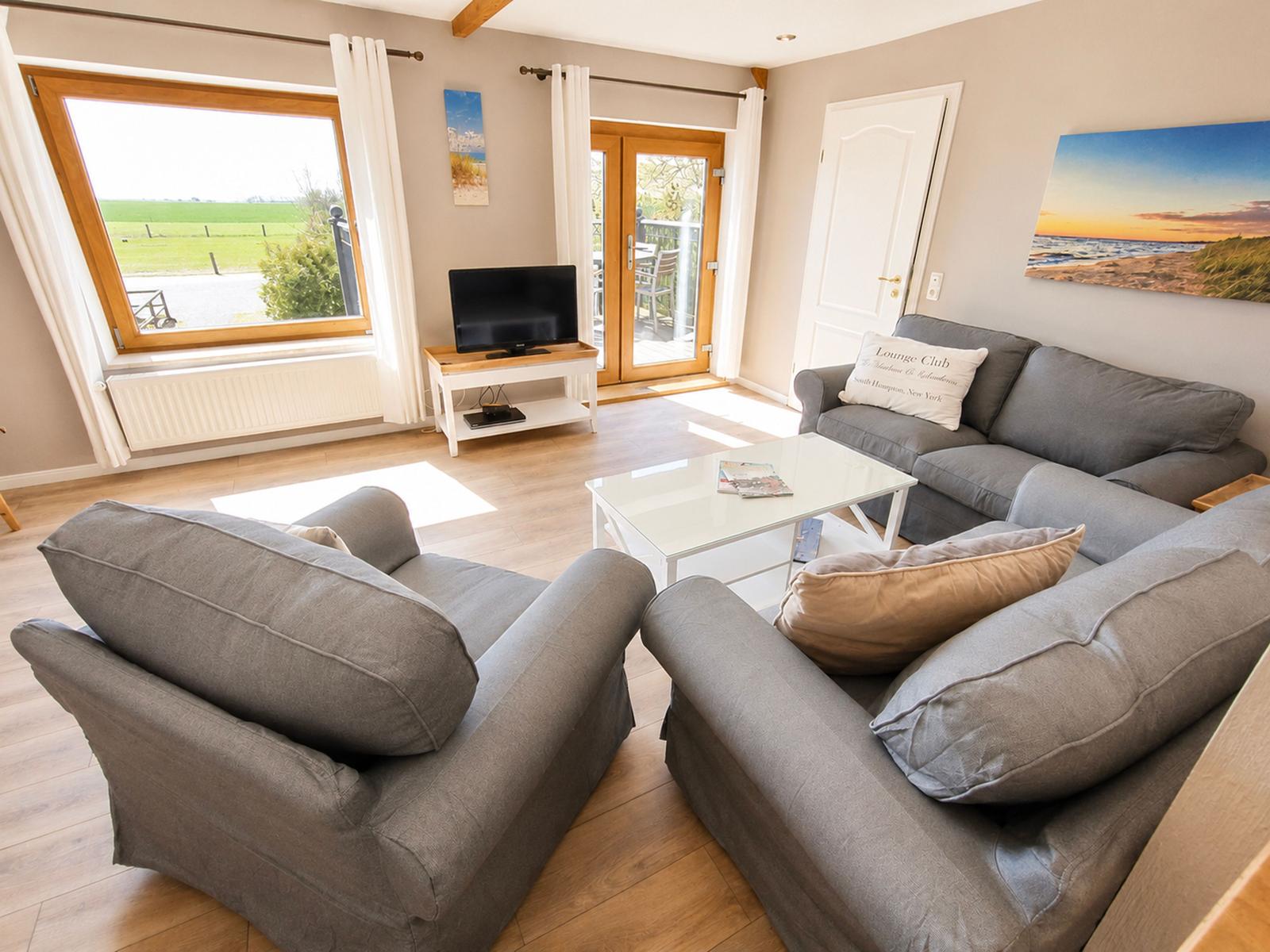 Wohnzimmer mit grauen Sitzmöbeln, Glas-Tisch und Fernseher. Durch Fenster und Tür Blick auf Garten.