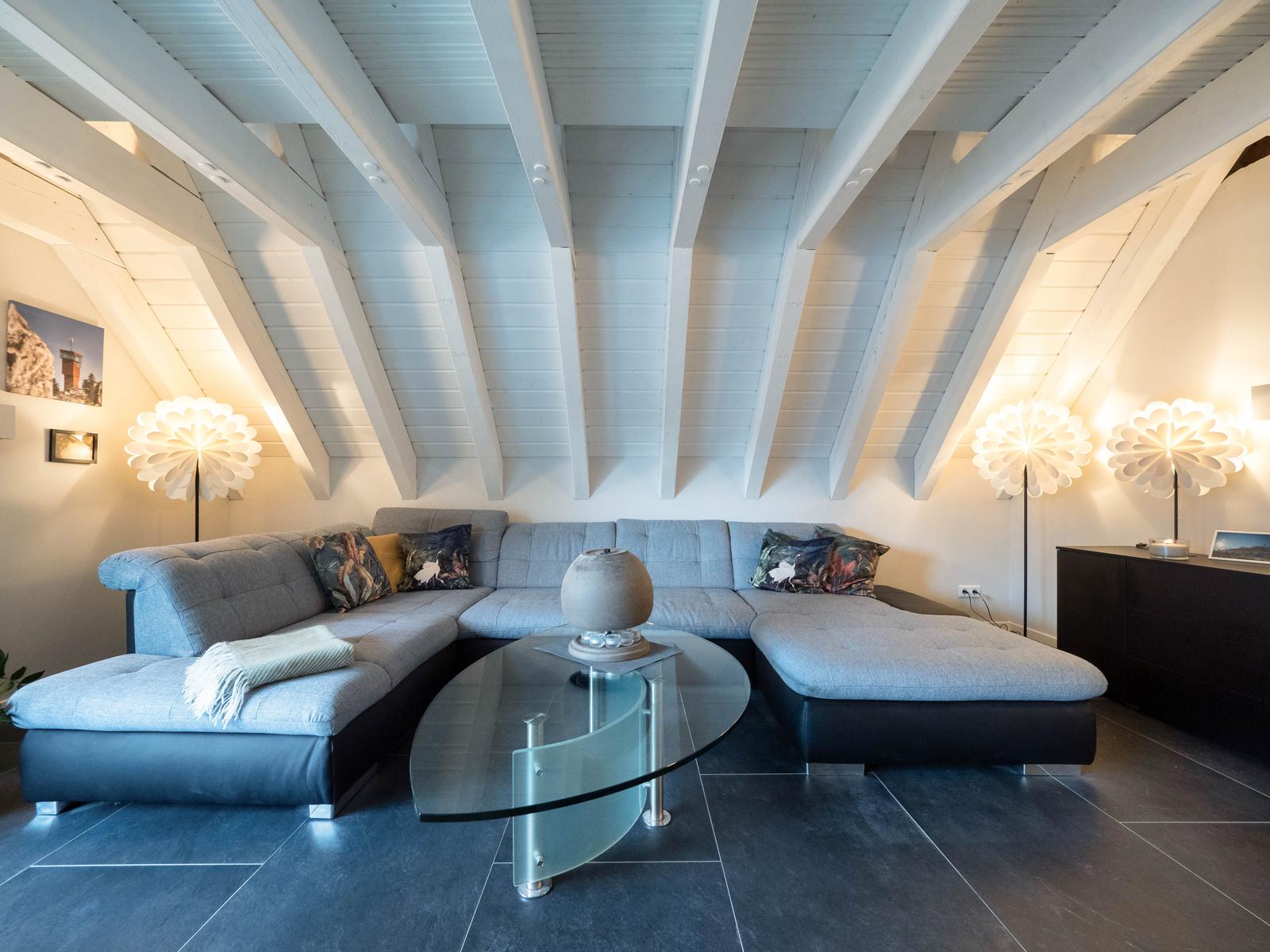 Wohnzimmer mit grauem Sofa, Glas-Tisch und weißen Dachschrägen.
