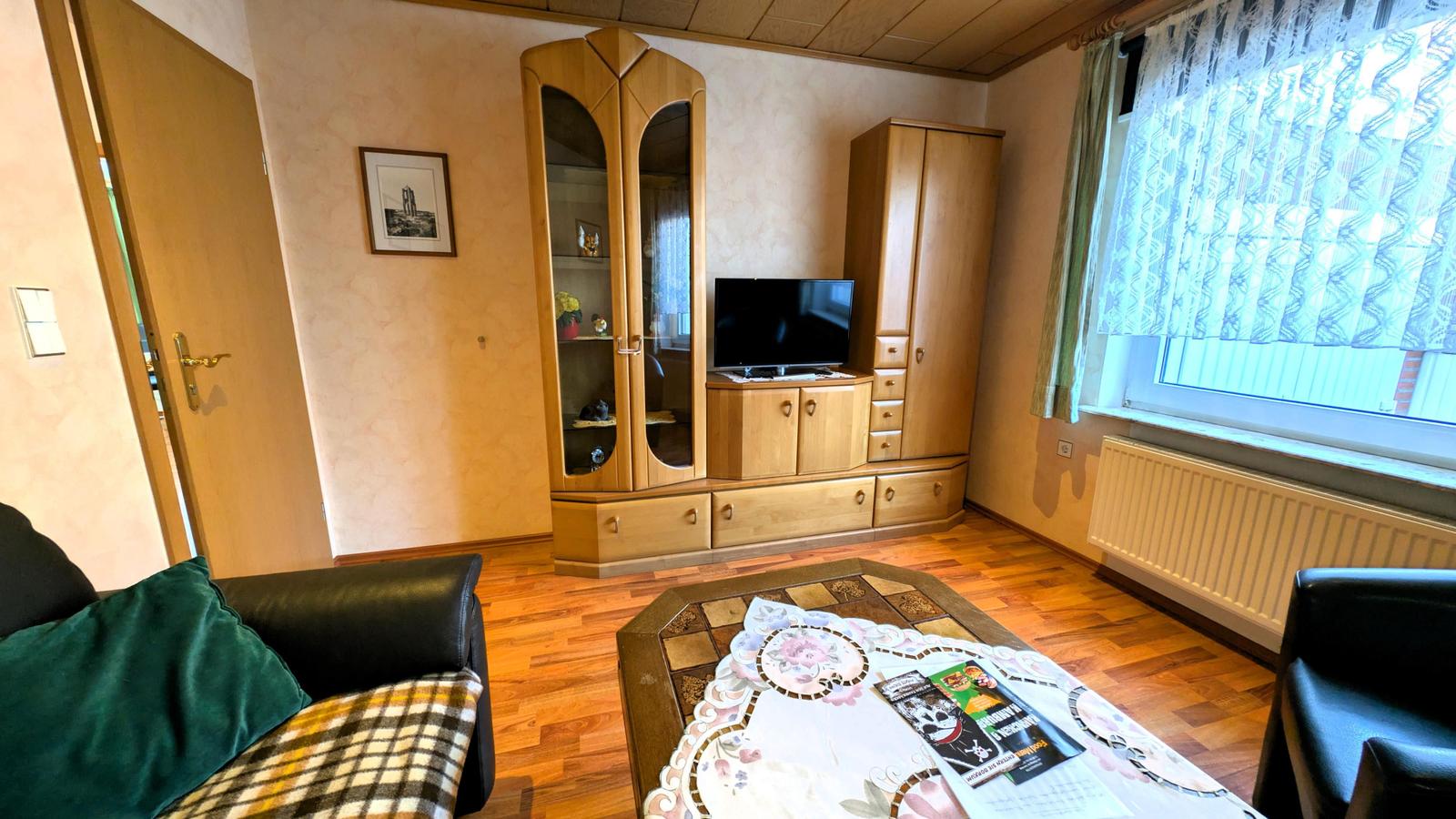 Wohnzimmer mit Holzmöbeln, Fernseher und Fenster mit Vorhängen.